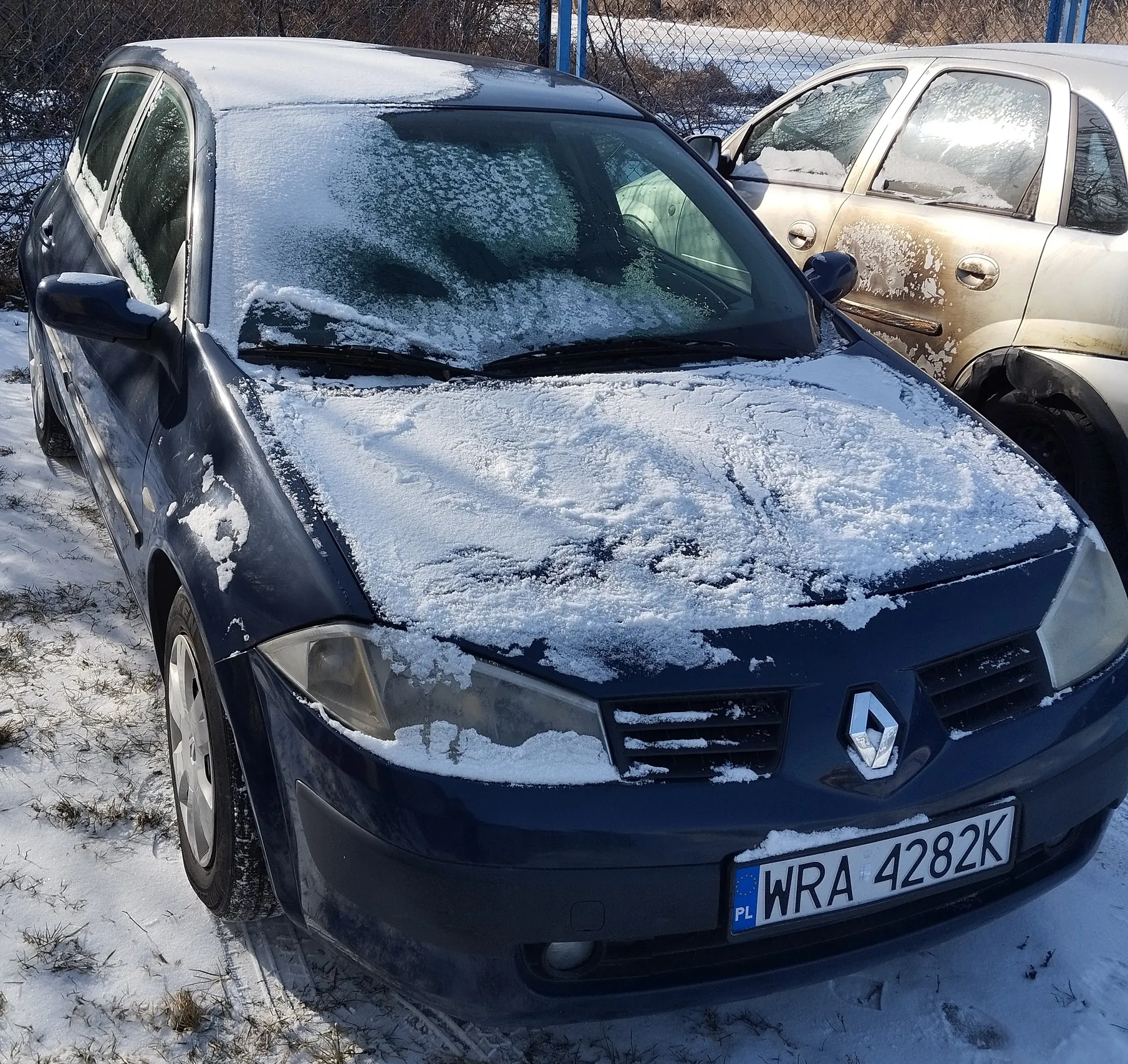 Ciemnoniebieski samochód marki Renault z tablicą rejestracyjną WRA 4282K (PL) pokryty śniegiem na masce, przedniej szybie i dachu. Obok stoi srebrny samochód również z śniegiem na karoserii. Tło: siatka ogrodzeniowa, śnieg na ziemi i suche trawy. Widoczny logo Renault na masce.