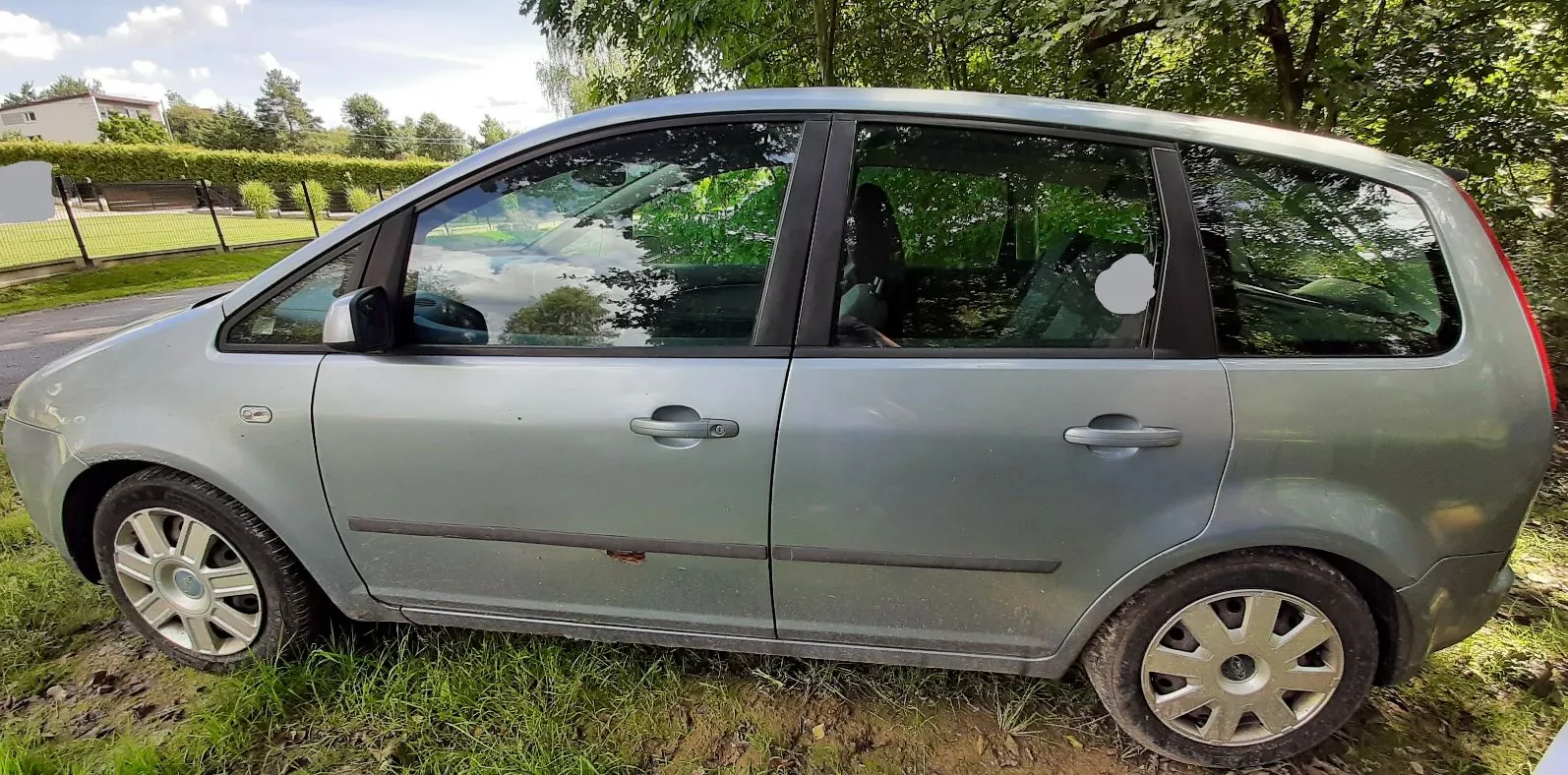 Boczny widok srebrno-szarego samochodu typu kombi zaparkowanego na trawie przed linią drzew. Pojazd jest widoczny z profilu, od przodu do tyłu.