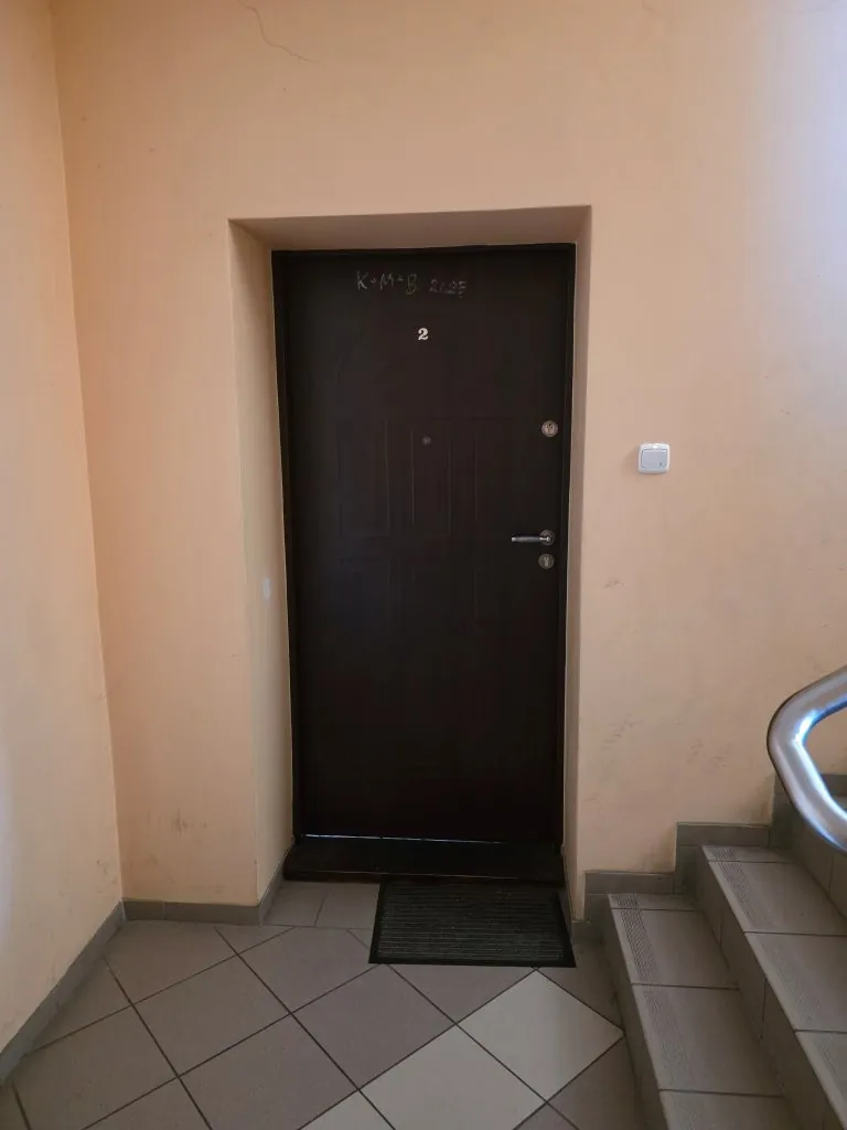 Ciemnobrązowe drzwi wejściowe w jasnej, beżowej ścianie. Nad drzwiami widnieje napis wykonany kredą: 'K+M+B 2023', a poniżej niego cyfra '2'. Przed drzwiami na szarej, płytkowej podłodze leży ciemna wycieraczka. Po prawej stronie drzwi znajduje się biały włącznik światła oraz fragment metalowej barierki schodów.