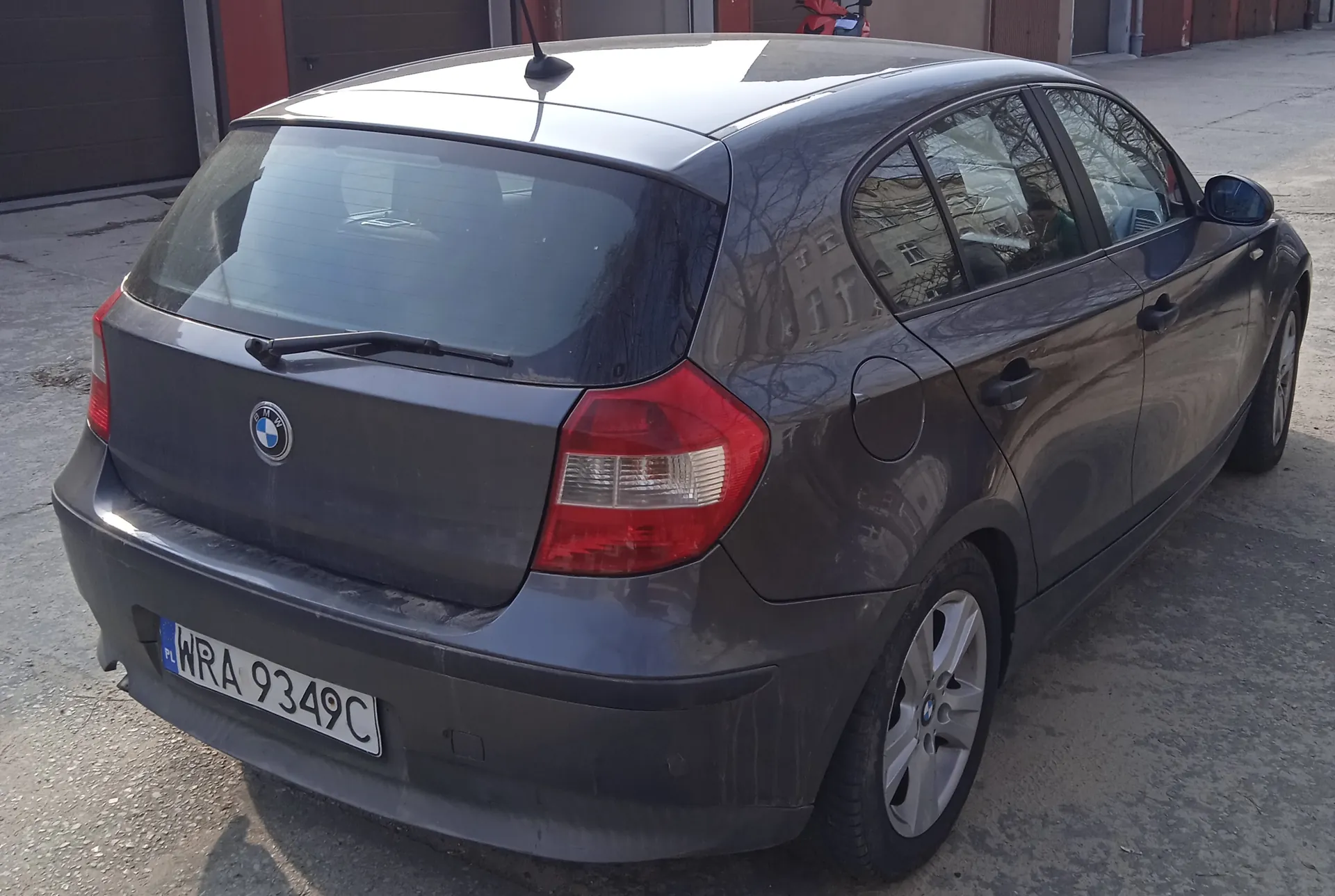 Szary samochód osobowy marki BMW typu hatchback zaparkowany na betonowej powierzchni. Widok z tyłu i boku. Na tylnej szybie widoczny wycieraczka, a na bagażniku logo BMW. Tablica rejestracyjna z białym tłem i czarnym napisem WRA 9349C z niebieskim pasem po lewej stronie z napisem PL. W tle zamknięte bramy garażowe, czerwony motocykl i budynki. Odbicia w szybach przedstawiają drzewa i budynki.