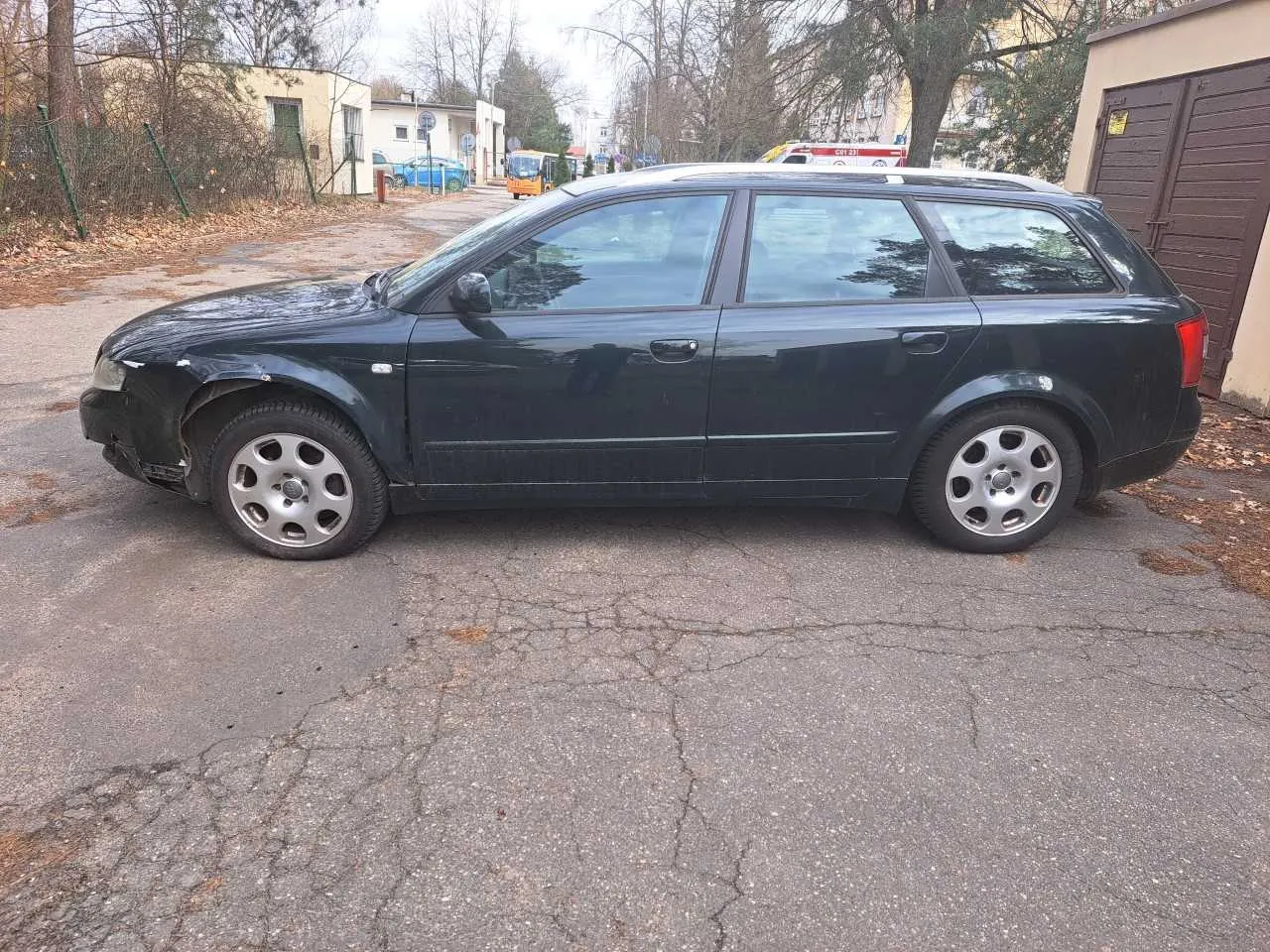 Ciemnozielony samochód kombi zaparkowany na pękniętej nawierzchni asfaltowej. Widoczne uszkodzenie przedniego lewego zderzaka. W tle budynki mieszkalne, drzewa bez liści, niebieski samochód, pomarańczowy pojazd i czerwono-biały ambulans. Na ziemi suche liście.