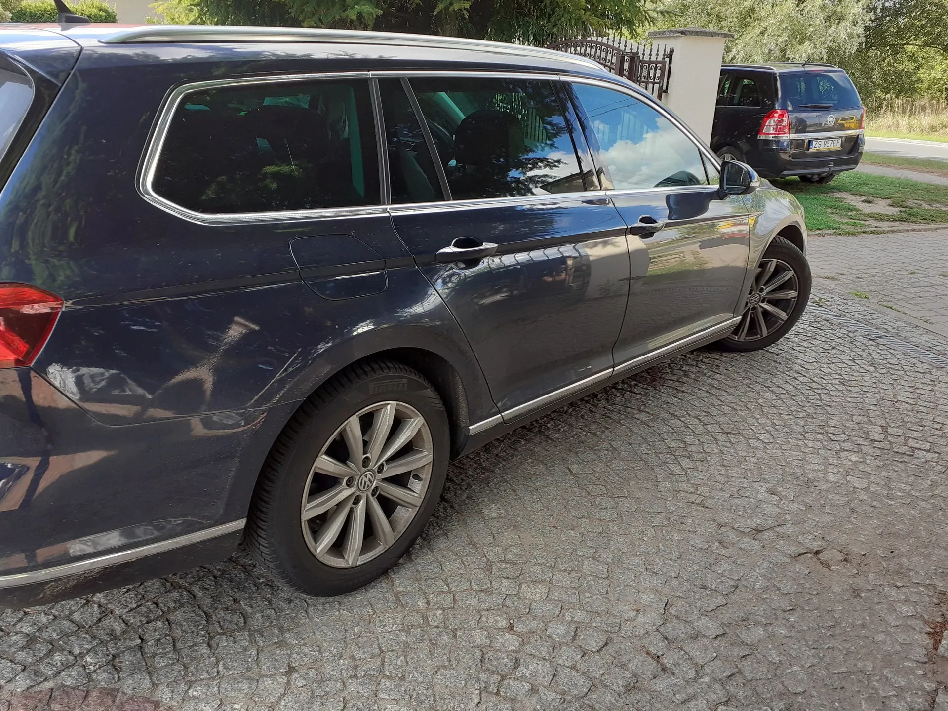 Boczny widok ciemnoniebieskiego samochodu typu kombi zaparkowanego na kostce brukowej. W tle widoczny jest drugi, czarny samochód oraz fragment ogrodzenia i zieleni.