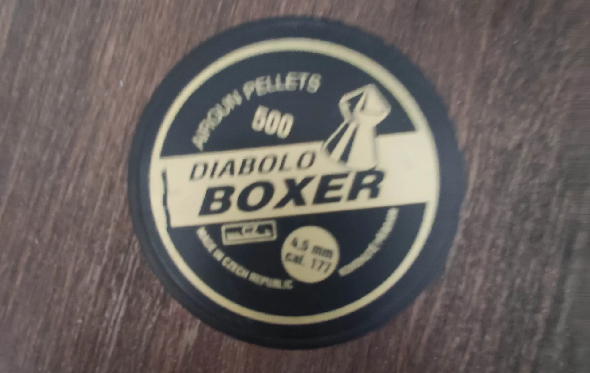 Okrągła, czarna nakrętka lub wieczko z żółtymi napisami. Na górze widnieje napis 'AIRGUN PELLETS', poniżej liczba '500', a następnie słowa 'DIABOLO BOXER'. Po prawej stronie znajduje się grafika przedstawiająca śrut do wiatrówki. Na dole widnieje napis 'MADE IN CZECH REPUBLIC' oraz oznaczenia kalibru: '4.5 mm Cal. 177'. Całość znajduje się na ciemnobrązowym, drewnianym tle.
