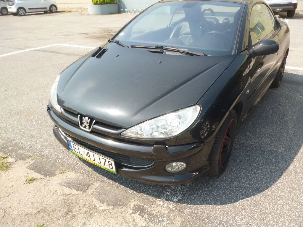 Czarny samochód osobowy marki Peugeot zaparkowany na asfaltowym placu. Widoczny jest przód pojazdu z tablicą rejestracyjną o numerze EL 4JJ78. W tle widać fragment innego, białego samochodu oraz betonowe krawężniki.