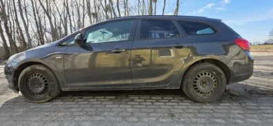 Ciemnoszary samochód typu kombi zaparkowany na brukowanej powierzchni. W tle wysokie, bezlistne drzewa i błękitne niebo z nielicznymi chmurami. Samochód ma zabrudzone koła i widoczne reflektory tylne.