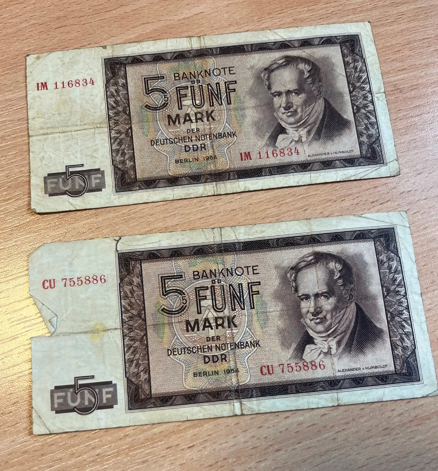 Dwa banknoty o nominale 5 marek z Niemieckiej Republiki Demokratycznej (DDR) z 1954 roku, leżące na drewnianej powierzchni. Na każdym z banknotów widnieje portret Aleksandra von Humboldta oraz napis 'BANKNOTE 5 FUNF MARK DER DEUTSCHEN NOTENBANK DDR BERLIN 1954'. Górny banknot ma numer seryjny IM 116834, a dolny CU 755886. Banknoty są w stanie zużytym, z widocznymi zagięciami i zabrudzeniami.