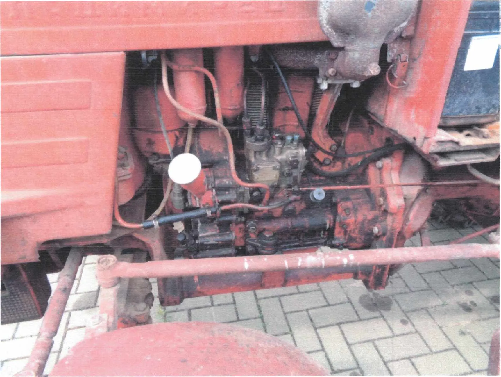 Zbliżenie na czerwony silnik mechaniczny z widocznymi elementami takimi jak rury, przewody, zawory i metalowe części. Silnik wykazuje oznaki zużycia i rdzy. W tle widoczna jest brukowana powierzchnia. Na silniku znajduje się biały okrągły element oraz czarne przewody.