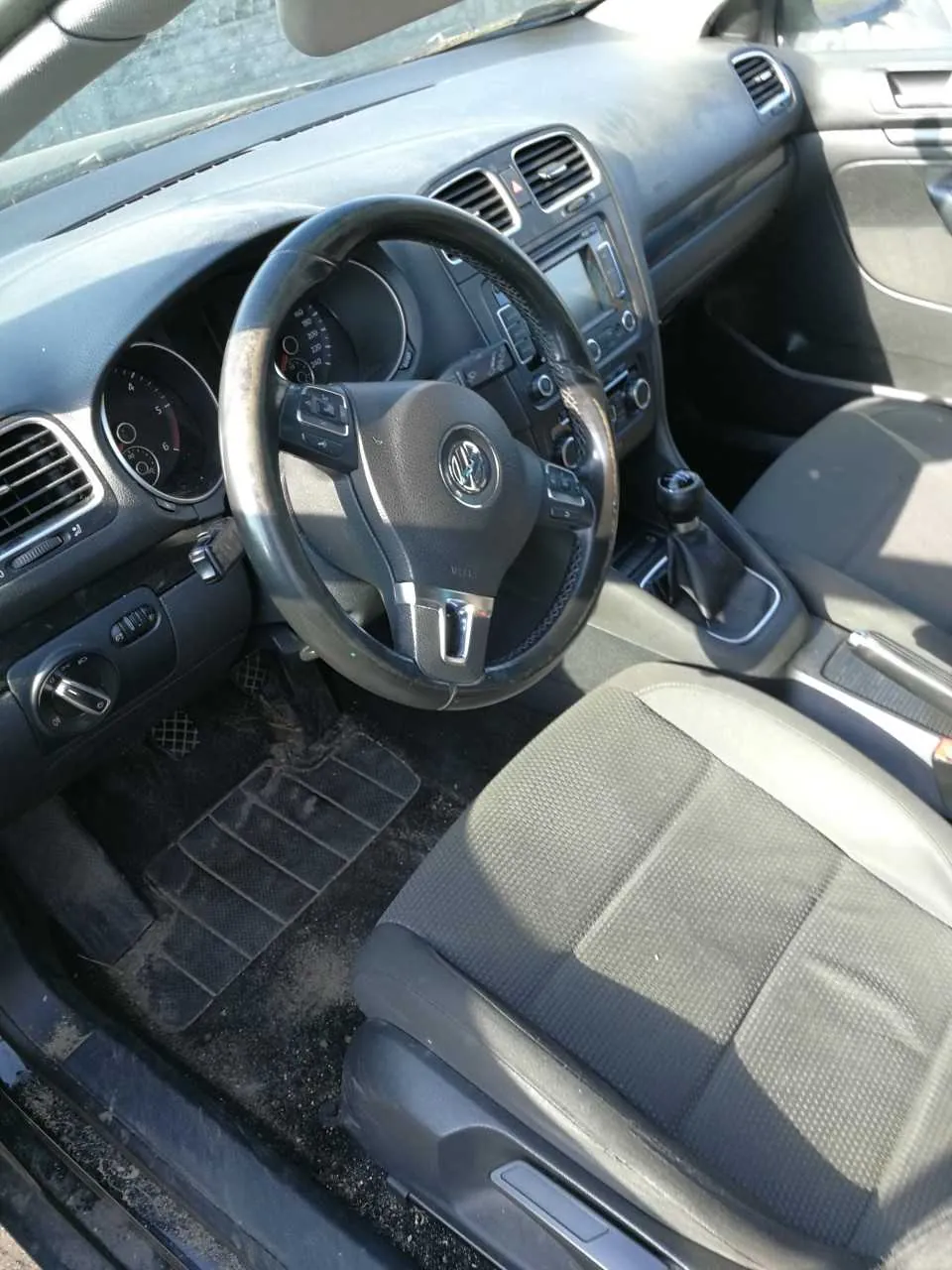 Wnętrze samochodu marki Volkswagen z widocznym kierownicą z logo VW, tablicą rozdzielczą z licznikami, przyciskami sterującymi, dźwignią zmiany biegów, siedzeniami oraz brudnymi dywanikami podłogowymi. W tle widoczne są wentylacyjne otwory i fragment przedniej szyby.