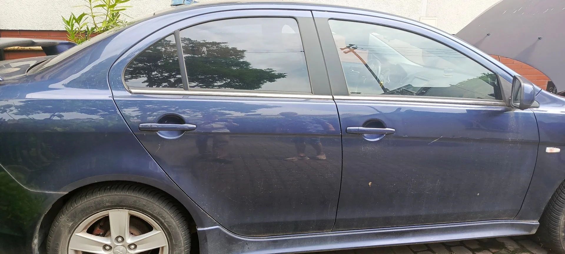 Boczny widok niebieskiego samochodu osobowego typu sedan. Widoczne są przednie i tylne drzwi z klamkami, szyby z odbiciem drzew oraz przednie i tylne koło z felgami aluminiowymi. Karoseria jest lekko zakurzona.