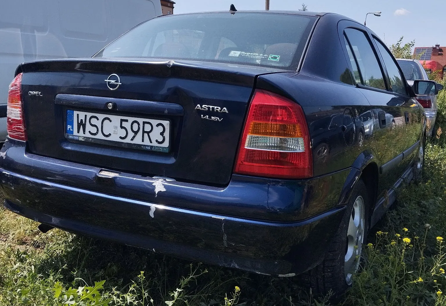 Tylny widok granatowego samochodu marki Opel Astra 1.4 16V. Pojazd posiada polską tablicę rejestracyjną o numerze WSC 59R3. Lakier na tylnym zderzaku jest uszkodzony i odpryskuje. Samochód stoi na trawie z żółtymi kwiatami.