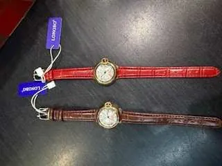 Dwa zegarki na rękę leżące na ciemnej powierzchni. Górny zegarek ma czerwony pasek imitujący skórę krokodyla i złotą obudowę, dolny brązowy pasek i srebrną obudowę. Obaj mają białe tarcze z czarnymi indeksami. Do każdego zegarka przyczepiona jest niebieska etykieta z białym tekstem: górna etykieta z napisem 'JORDAN', dolna z napisem 'JORDAN'S'.