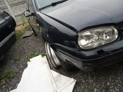 Ciemny samochód osobowy z widocznym uszkodzeniem przedniej części nadwozia, zaparkowany na podłożu z żwiru. W pierwszym planie leżą białe arkusze materiału. Po lewej stronie częściowo widoczny inny samochód, w tle ceglana ściana i fragment trawy. Samochód ma srebrne felgi i reflektory z przodu.