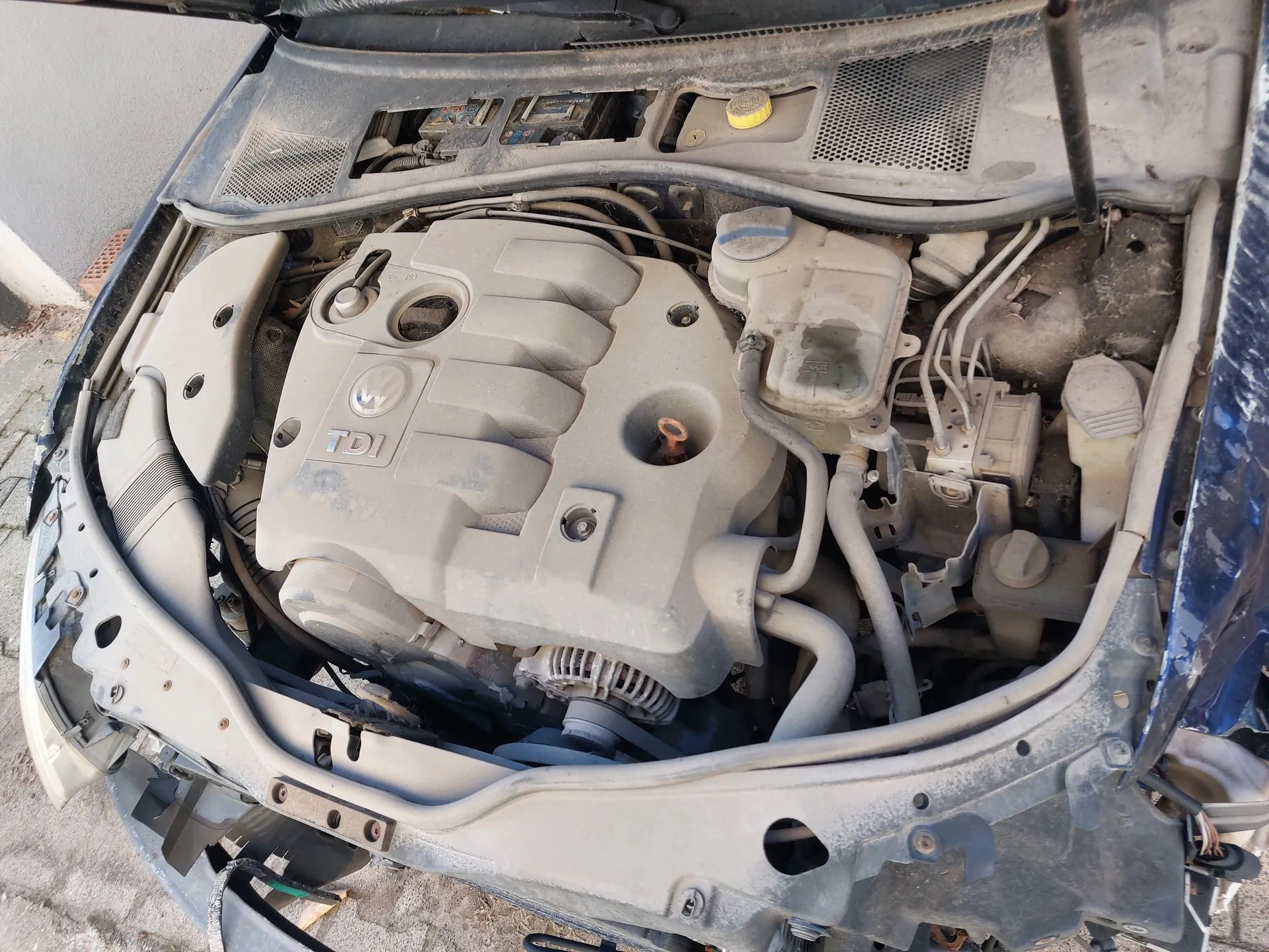 Widok z góry na zakurzoną komorę silnika w niebieskim samochodzie. Centralnie znajduje się plastikowa osłona silnika z logo Volkswagena oraz napisem 'TDI'. Po prawej stronie widoczne są zbiorniki z płynami eksploatacyjnymi, przewody i elementy mechaniczne. Całość jest pokryta warstwą szarego pyłu.