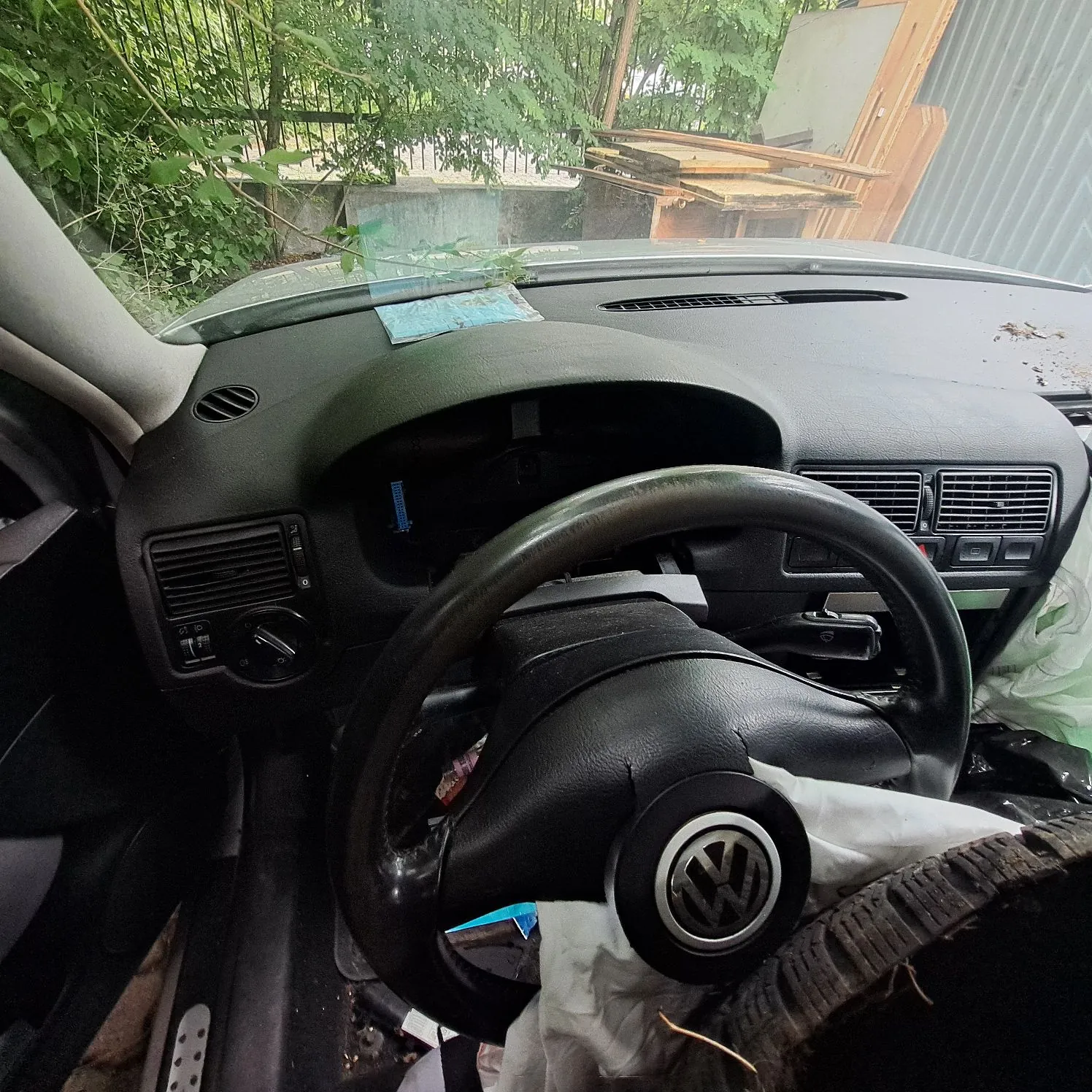 Wnętrze samochodu marki Volkswagen, widok z perspektywy kierowcy. Widoczna jest czarna kierownica z logo VW w centrum, deska rozdzielcza z pustym miejscem po liczniku oraz kratki nawiewów. Wewnątrz auta panuje nieład, na siedziach i podłodze znajdują się białe folie oraz inne przedmioty. Przez przednią szybę widoczna jest zieleń roślin i fragmenty drewnianych konstrukcji.
