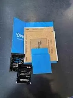 Na ciemnej powierzchni leżą różne przedmioty: niebieski folder z częściowo widocznym napisem "Digi", brązowa koperta z tekstem, niebieski prostokątny element oraz dwie czarne karty z napisami, w tym jedna z widocznym logo "Digi". Wszystkie elementy są ułożone płasko, bez widocznych akcji lub ruchu.