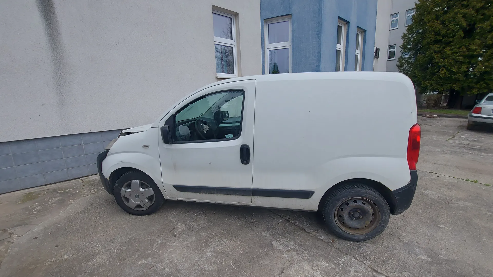 Biały furgon zaparkowany na betonowej powierzchni przed budynkiem o szarej i niebieskiej elewacji. Furgon ma uszkodzony przód, brudne boki i zardzewiałe tylne koło. W tle widoczne są okna z białymi ramami, drzewo oraz częściowo inny samochód. Betonowa powierzchnia posiada pęknięcia i plamy.