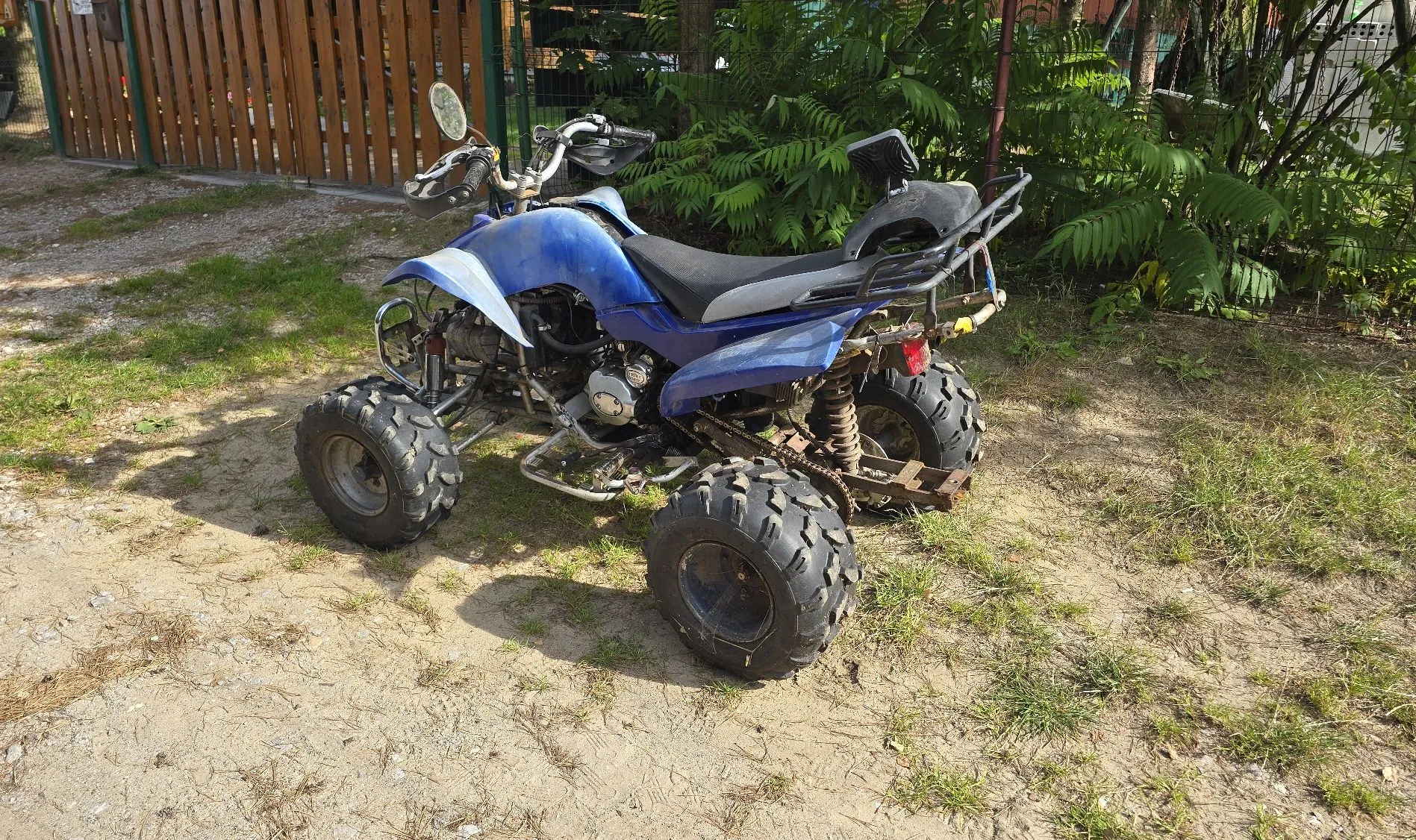 Niebieski quad zaparkowany na piaszczysto-trawiastej nawierzchni. Pojazd ma duże, terenowe opony, czarne siedzenie i metalowy bagażnik z tyłu. W tle widoczny jest drewniany płot oraz zielone krzewy i roślinność.