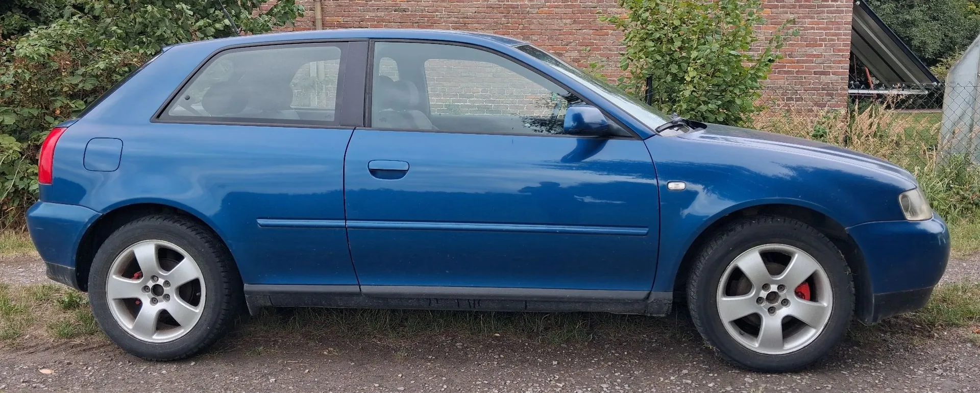 Niebieski samochód osobowy typu hatchback zaparkowany bokiem na żwirowej nawierzchni na tle ceglanego muru i zielonych krzewów.