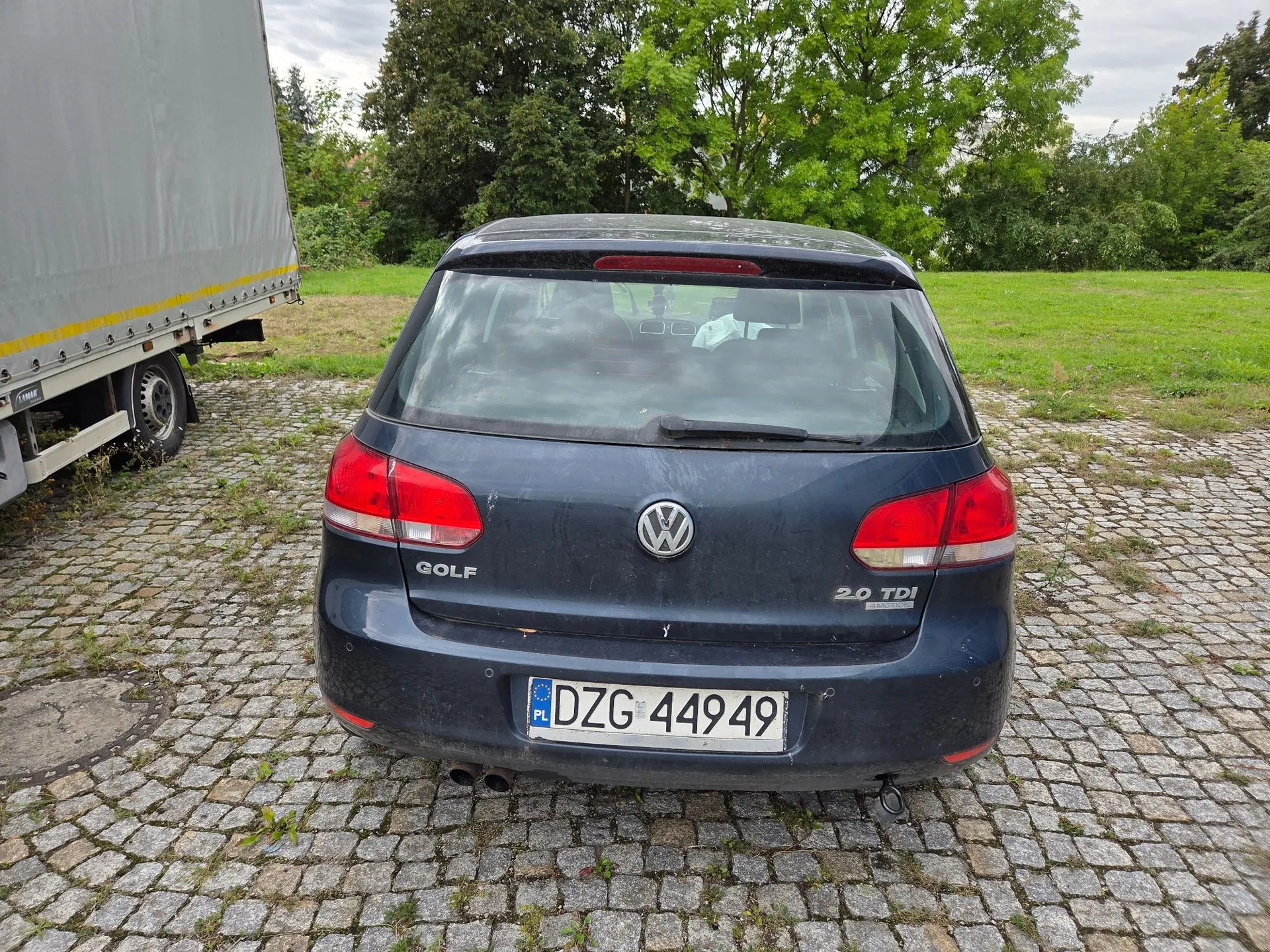 Tylny widok ciemnoniebieskiego samochodu Volkswagen Golf zaparkowanego na kostce brukowej. Na klapie bagażnika widoczne jest logo Volkswagena, napis GOLF po lewej stronie oraz oznaczenie 2.0 TDI po prawej. Samochód posiada polską tablicę rejestracyjną o numerze DZG 44949. Po lewej stronie kadru widoczny jest fragment białej naczepy ciężarówki. W tle znajduje się zielona trawa i drzewa.