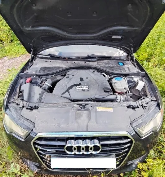 Widok z przodu na czarny samochód marki Audi z otwartą maską, odsłaniającą silnik. Na osłonie silnika widoczne jest logo Audi oraz napis 'V6 TDI'. Samochód stoi na trawiastym podłożu.