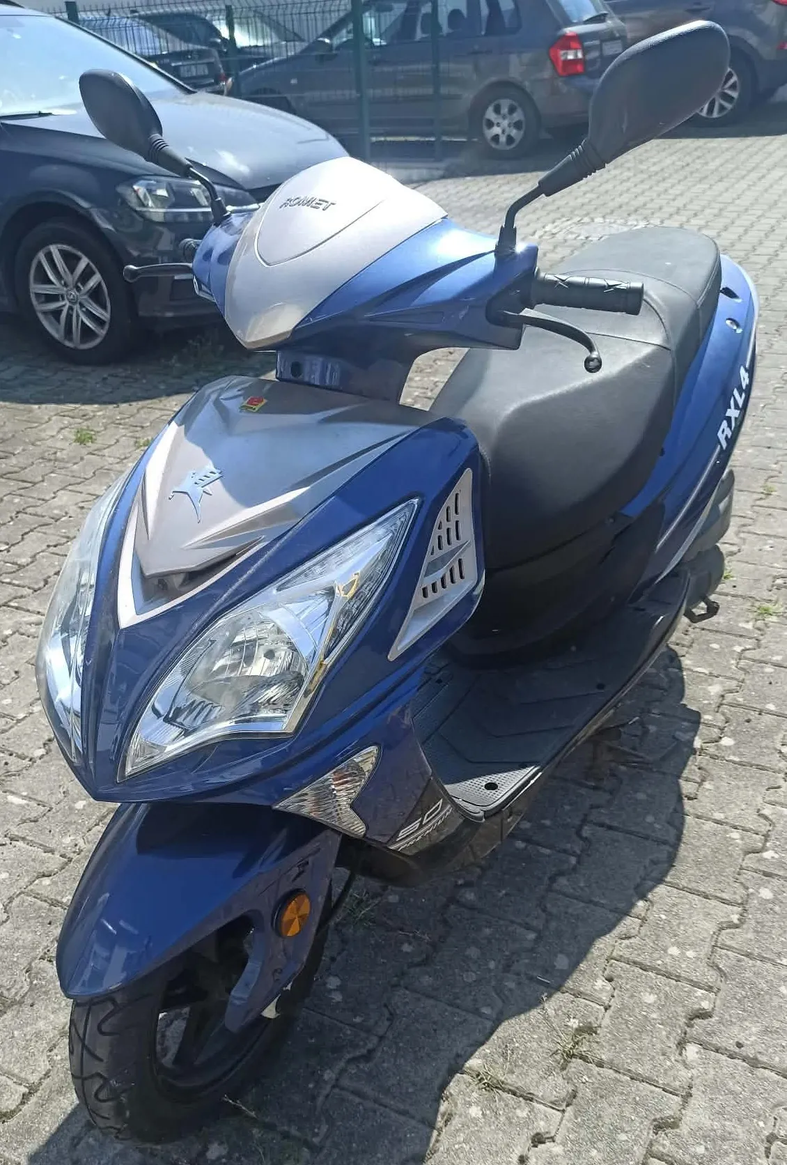 Niebieski skuter marki Romet, model RXL, zaparkowany na kostce brukowej. Skuter jest ustawiony przodem do widza, widoczny jest reflektor, przednia owiewka z logo oraz siedzenie. W tle widać zaparkowane samochody i ogrodzenie z siatki.