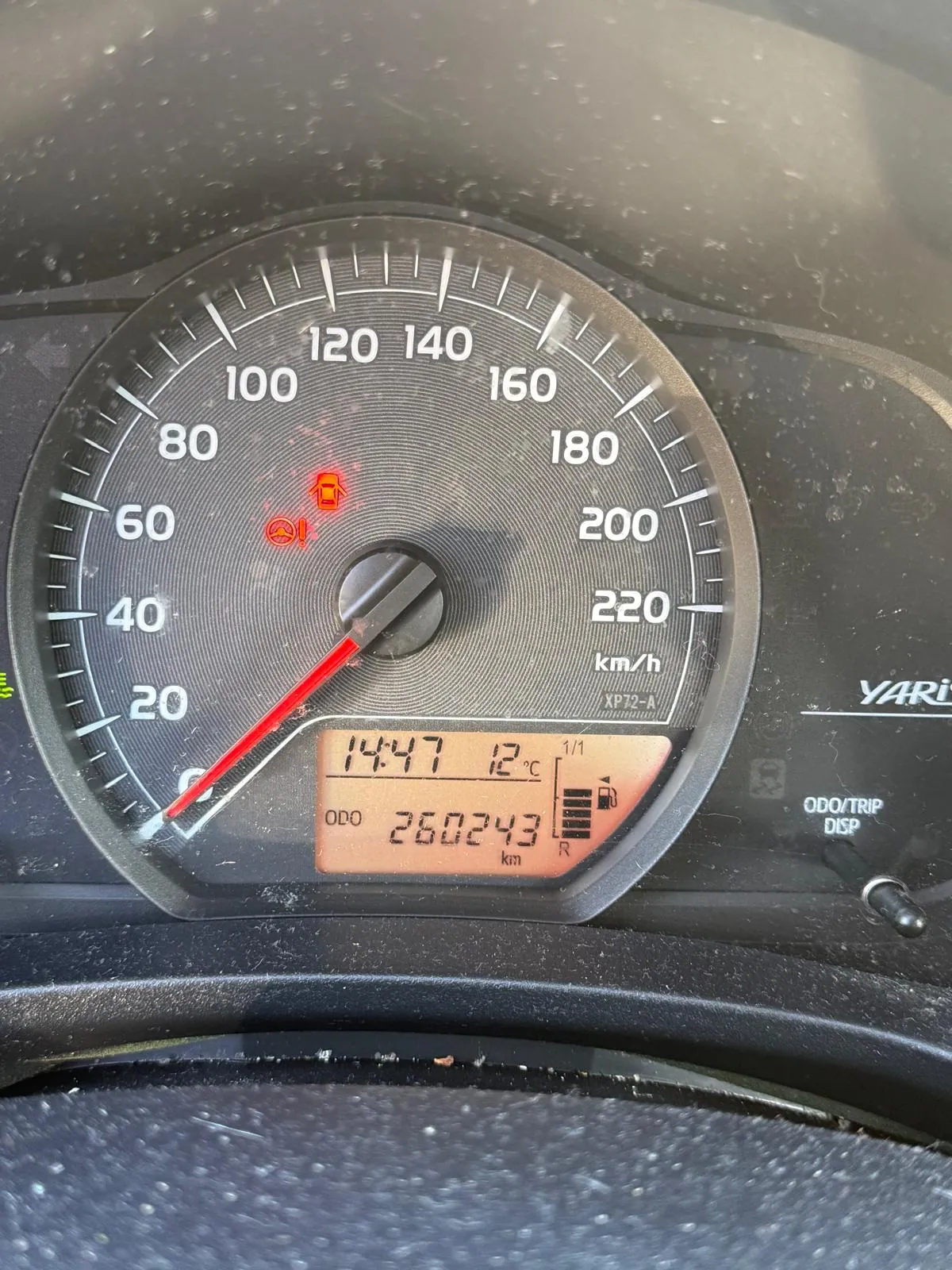 Zbliżenie na zegar samochodowy marki Toyota Yaris. Wskazówka prędkościomierza wskazuje 0 km/h. Na wyświetlaczu cyfrowym widoczna godzina 14:47, temperatura 12°C oraz przebieg 260243 km. Aktywne są dwa czerwone sygnalizatory: ikona samochodu z wykrzyknikiem oraz symbol oleju. Obok prędkościomierza umieszczono napisy 'YARIS', 'ODO/TRIP DISP' oraz wskaźnik paliwa. Tło zegara ma czarny kolor z białymi oznaczeniami prędkości od 0 do 220 km/h.