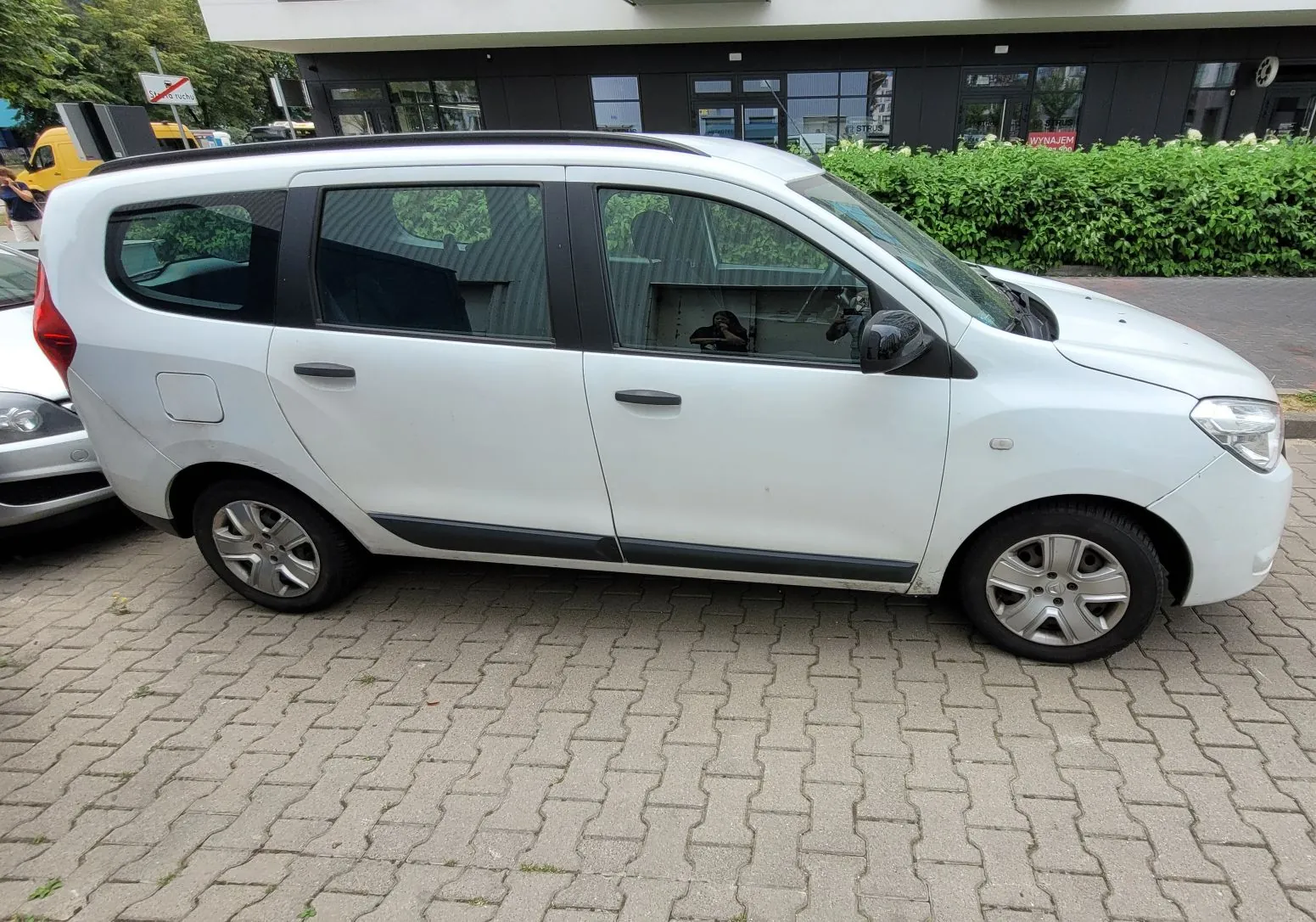 Biały samochód typu minivan zaparkowany bokiem na kostce brukowej przed nowoczesnym, ciemnym budynkiem z dużymi przeszkleniami i zielonym żywopłotem w tle.