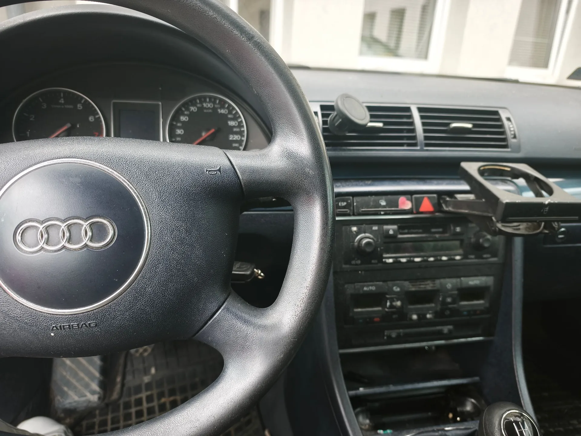 Wnętrze samochodu marki Audi widziane z perspektywy kierowcy. Na pierwszym planie znajduje się czarna, skórzana kierownica z logo Audi i napisem AIRBAG. W tle widoczna jest deska rozdzielcza z zegarami (obrotomierzem i prędkościomierzem), radiem, panelem klimatyzacji oraz uchwytem na telefon zamontowanym po prawej stronie.