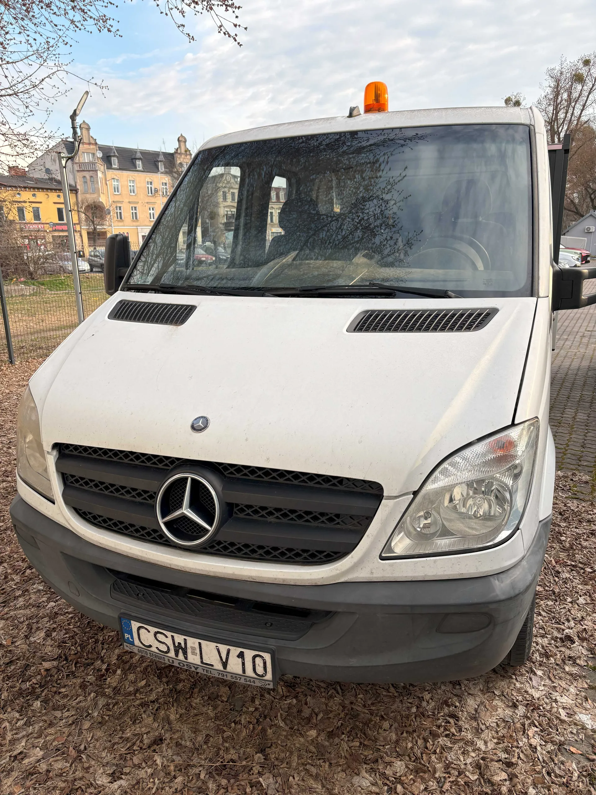 Biały samochód dostawczy marki Mercedes-Benz z widocznym logo na masce i tablicą rejestracyjną CSW LV10 z kodem PL. Na dachu pojazdu znajduje się pomarańczowy sygnalizator. W tle budynki mieszkalne, ogrodzenie i suche liście na ziemi. Niebo częściowo zachmurzone.