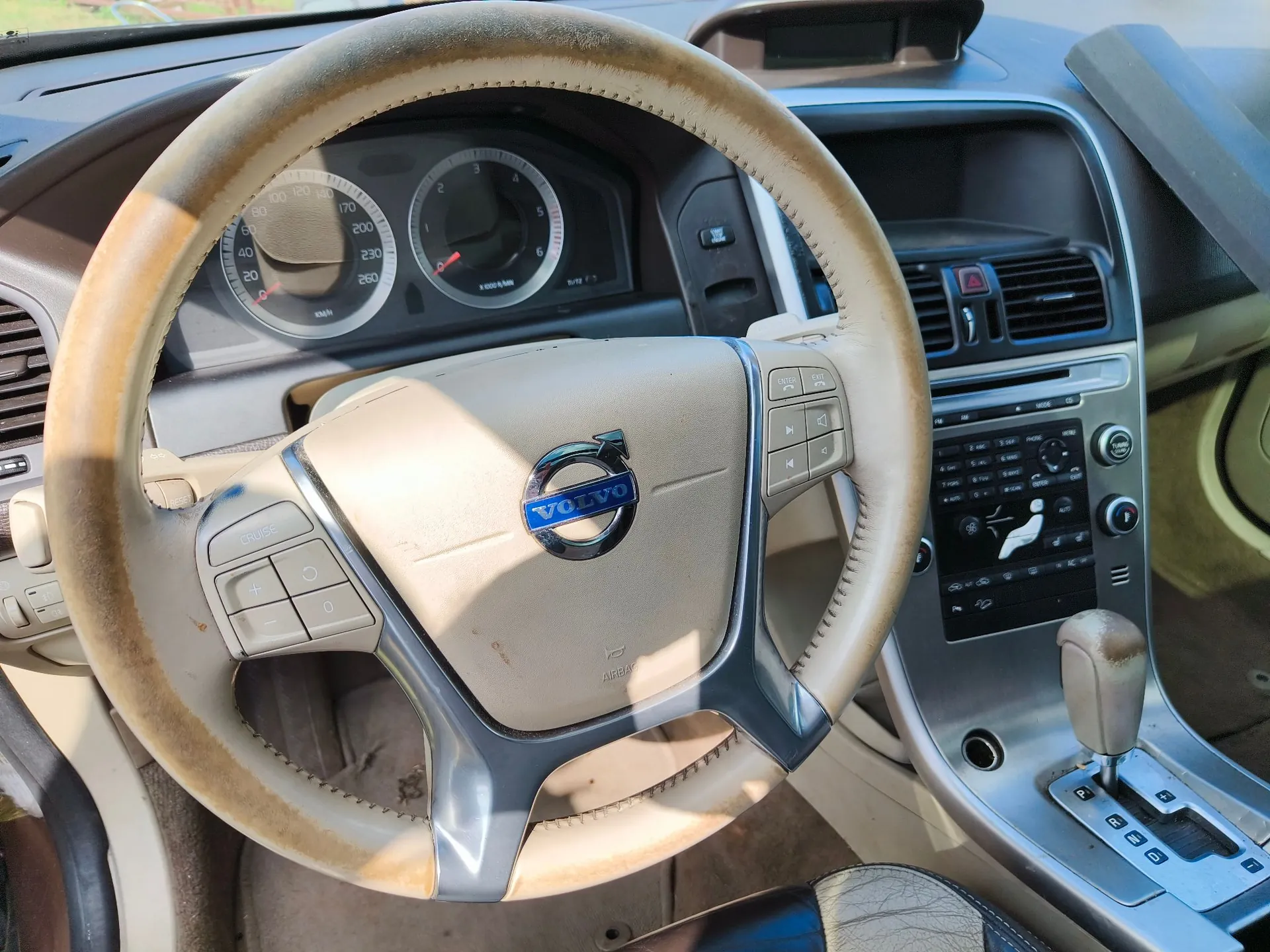 Wnętrze samochodu marki Volvo, widok z perspektywy kierowcy. W centrum znajduje się brązowa, skórzana kierownica z logo Volvo na środku. W tle widoczna jest deska rozdzielcza z analogowymi wskaźnikami prędkości i obrotów oraz konsola środkowa z systemem audio i lewarkiem zmiany biegów.
