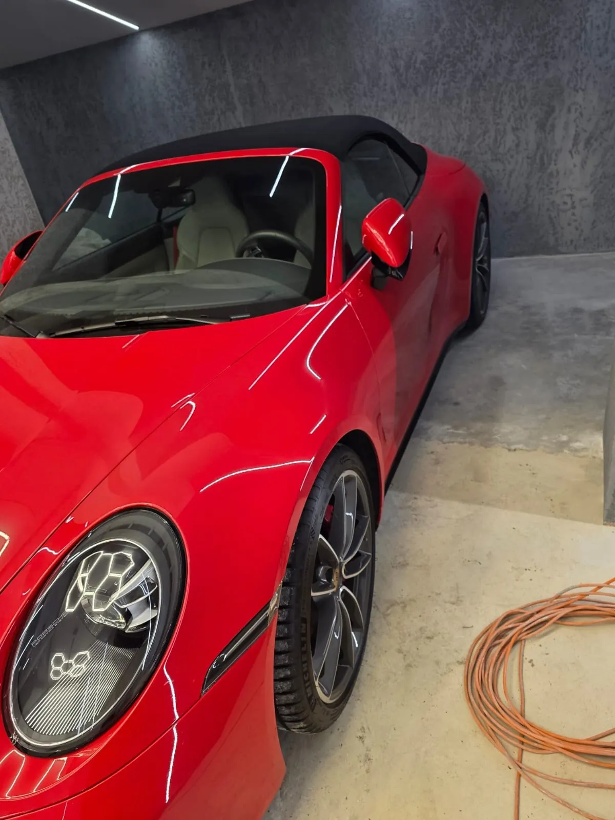 Czerwony samochód kabriolet z czarnym dachem, zaparkowany w garażu. Widoczne są białe siedzenia, kierownica, reflektory z geometrycznym wzorem oraz pomarańczowy przewód zwinięty na betonowej podłodze. Tło stanowi szara, teksturowana ściana i sufit z oświetleniem.