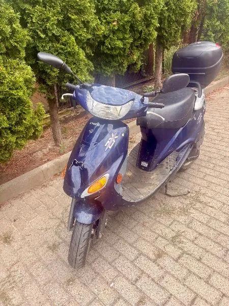 Granatowy skuter zaparkowany na kostce brukowej przed zielonymi krzewami. Pojazd posiada czarną kanapę, czarny kufer z tyłu oraz przednią lampę. Na przedniej owiewce widnieje napis 'Kyugo'.