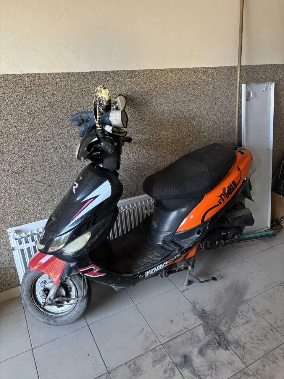 Czarno-pomarańczowy skuter zaparkowany w pomieszczeniu. Skuter ma czarne siedzenie, widoczne napisy 'Diablo' na pomarańczowej części oraz 'TORO' na czarnej części. Na kierownicy znajduje się zniszczony kask. W tle: ściana z dwoma rodzajami wykończenia (górna część gładka, beżowa; dolna część kruszona, brązowa), biały grzejnik i biały panel oparty o ścianę. Podłoga z szarymi płytkami z widocznymi plamami brudu.