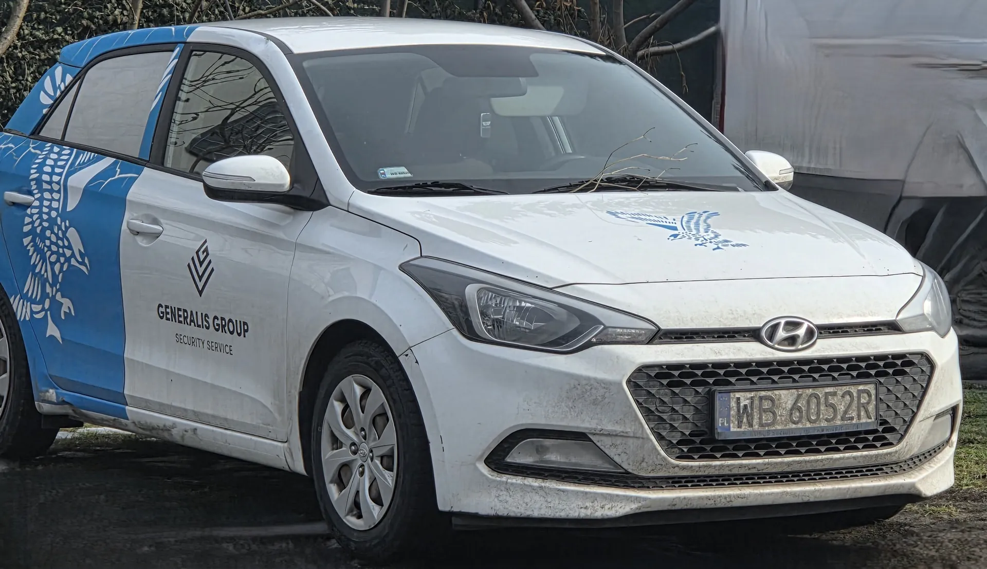Biały samochód marki Hyundai z niebieskimi akcentami, na boku widoczny duży stylizowany orzeł oraz napisy 'GENERALIS GROUP' i 'SECURITY SERVICE'. Na masce znajduje się niebieski motyw orła, a na przodzie tablica rejestracyjna 'WB 6052R' z oznaczeniem 'PL'. Samochód jest zabrudzony, stoi na nawierzchni z trawą w tle oraz zasłoniętym obiektem po prawej stronie. W tle widoczne są suche gałęzie drzew.