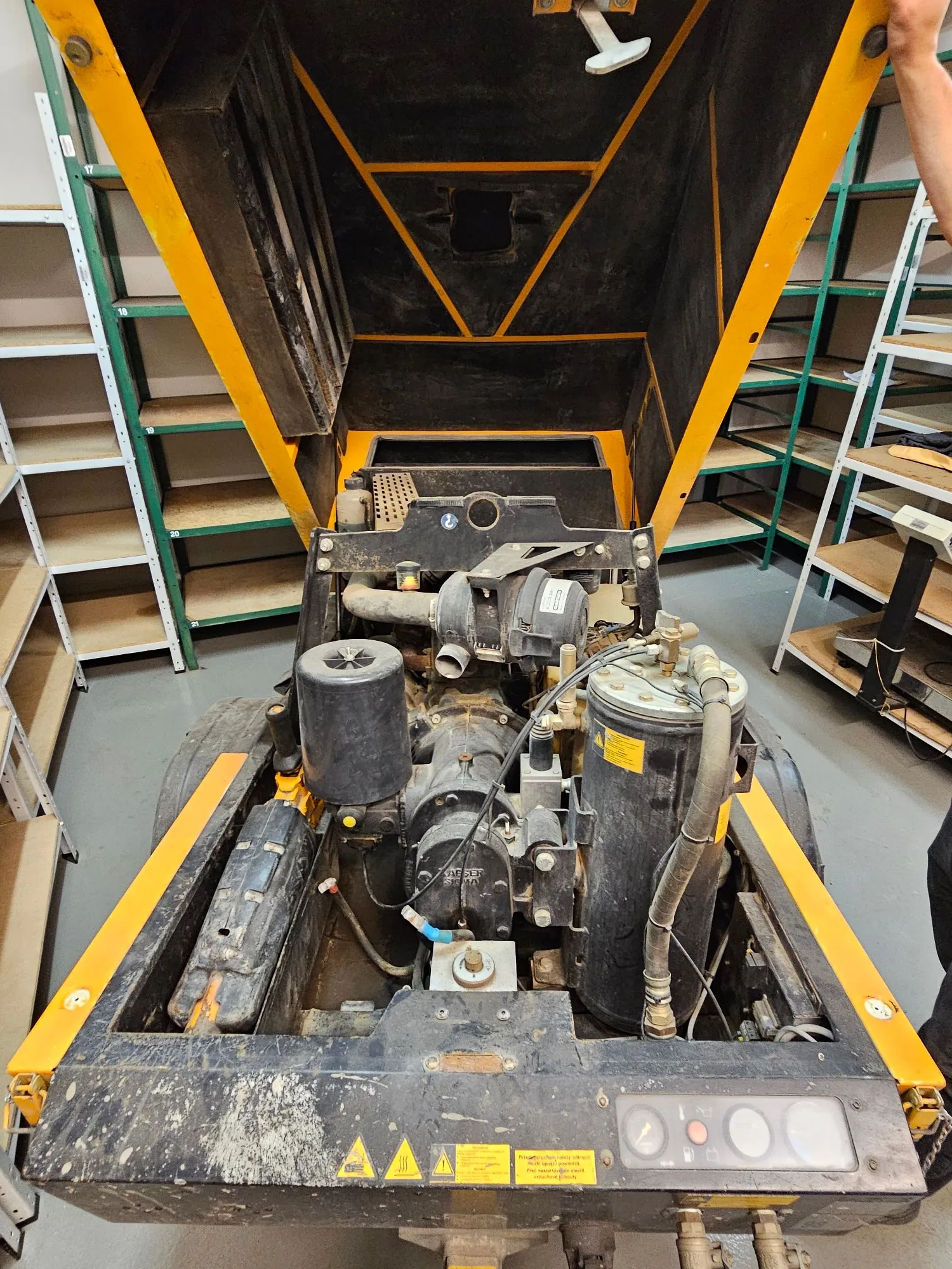 Zdjęcie przedstawia silnik i podzespoły mechaniczne maszyny przemysłowej w kolorach żółtym i czarnym. Widoczne są filtry oleju i paliwa, przewody oraz elementy konstrukcyjne. Po prawej stronie znajduje się panel z lampkami kontrolnymi i żółtą tabliczką ostrzegawczą. Obraz jest zorientowany poziomo, podczas gdy maszyna i tekst na tabliczkach są ustawione pionowo.