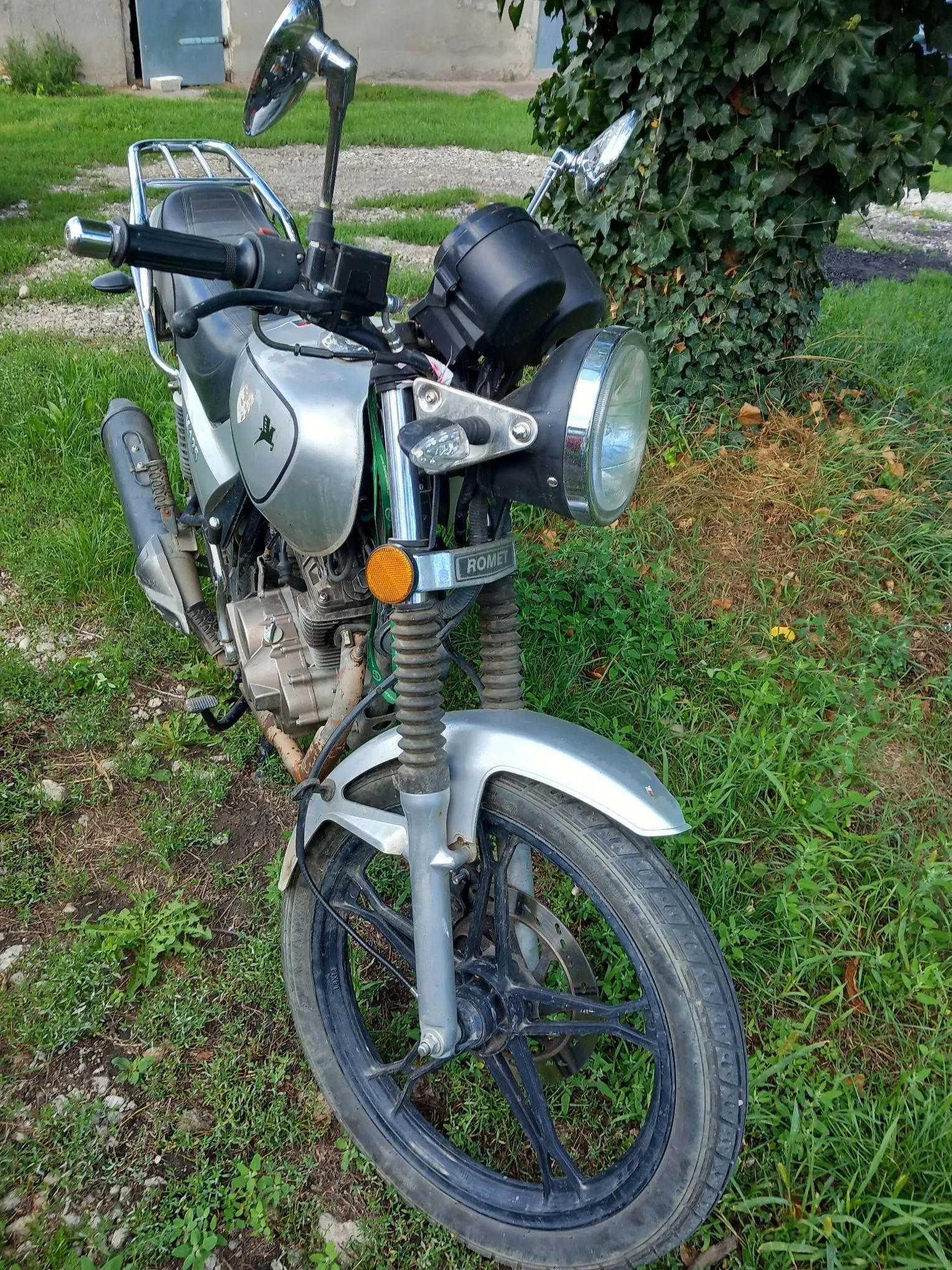 Srebrny motocykl marki Romet zaparkowany na trawie. Widoczny jest przód pojazdu z okrągłym reflektorem, przednim kołem z czarną felgą, kierownicą z lusterkami oraz srebrnym błotnikiem. Na przedniej części obudowy znajduje się niebieska tabliczka z białym napisem ROMET.