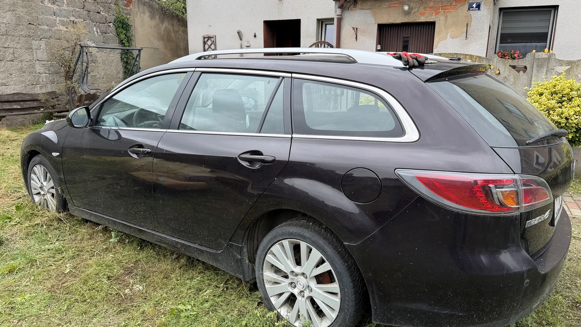 Czarny samochód typu kombi zaparkowany na trawiastym terenie. Pojazd jest widoczny z profilu od strony pasażera, z tyłu znajduje się biały budynek z oknami i drzwiami. Samochód ma srebrne felgi aluminiowe i srebrne relingi dachowe.