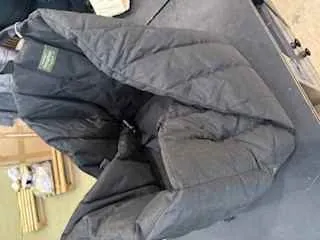 Ciemnoszara pikowana kurtka leżąca skręcona na powierzchni. Widoczny zielony etykietka z niewyraźnym tekstem na wewnętrznej stronie kurtki. W tle fragmenty drewnianych prętów i jasnej ściany. Obok kurtki znajduje się biały przedmiot oraz czarna powierzchnia z metalowymi elementami.