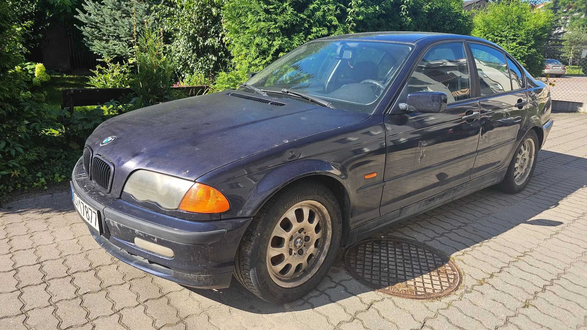 Ciemnoniebieski samochód osobowy marki BMW serii 3 zaparkowany na kostce brukowej. Pojazd jest zakurzony, z widocznymi śladami zużycia. W tle znajduje się gęsta, zielona roślinność i krzewy. Na ziemi obok samochodu widoczny jest okrągły właz kanalizacyjny.