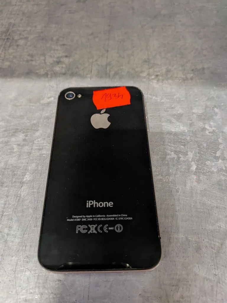 Tył czarnego smartfona iPhone leżącego na szarej, metalicznej powierzchni. Na górze obudowy znajduje się aparat fotograficzny oraz czerwona naklejka z ręcznym napisem. W centralnej części widoczne jest srebrne logo Apple, a poniżej napis 'iPhone'. Na dole znajduje się tekst: 'Designed by Apple in California Assembled in China Model A1367 EMC 2430 FCC ID: BCG-E2430A IC: 579C-E2430A' oraz znaki certyfikacji FC, CE i inne.