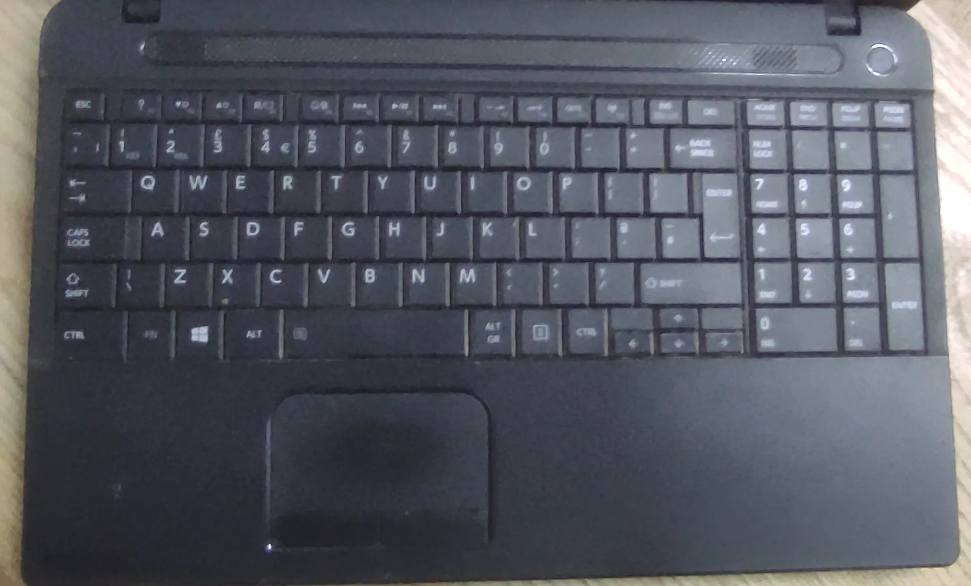 Widok z góry na czarną klawiaturę laptopa z gładzikiem poniżej. Klawiatura posiada standardowy układ QWERTY oraz blok numeryczny po prawej stronie.