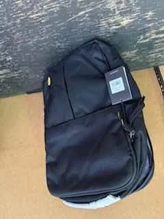 Czarny plecak z przodem z kieszenią na zamku, małym żółtym elementem i prostokątną etykietą. Plecak leży na powierzchni z ciemnego drewna i jasnobrązowym tłem.