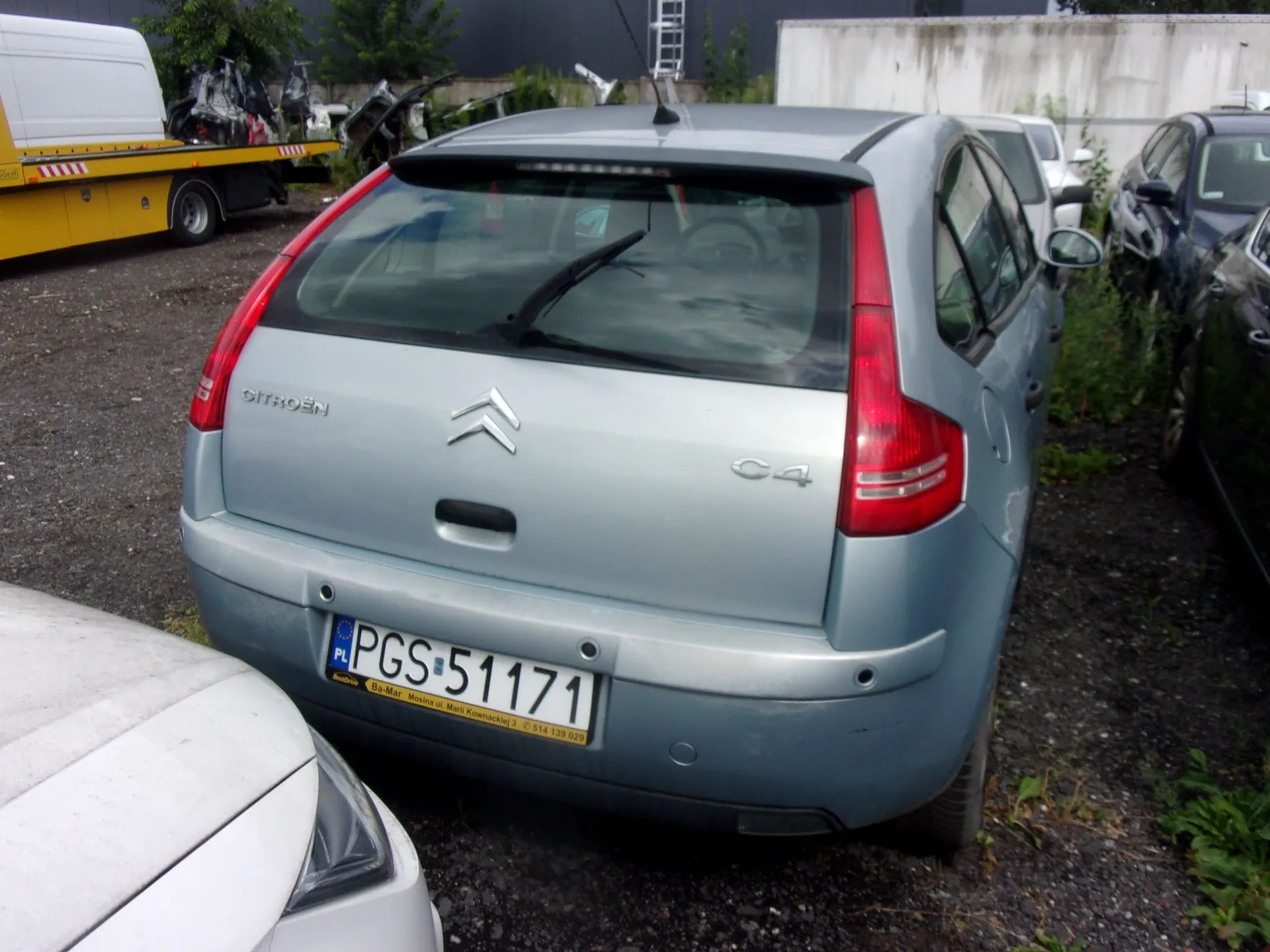 Tylny widok jasnoniebieskiego samochodu marki Citroën C4 zaparkowanego na żwirowym placu. Na klapie bagażnika widoczne są napisy 'CITROËN' oraz 'C4' oraz logo marki. Samochód posiada polską tablicę rejestracyjną o numerze PGS 51171. W tle widoczny jest żółty samochód dostawczy oraz inne zaparkowane pojazdy.