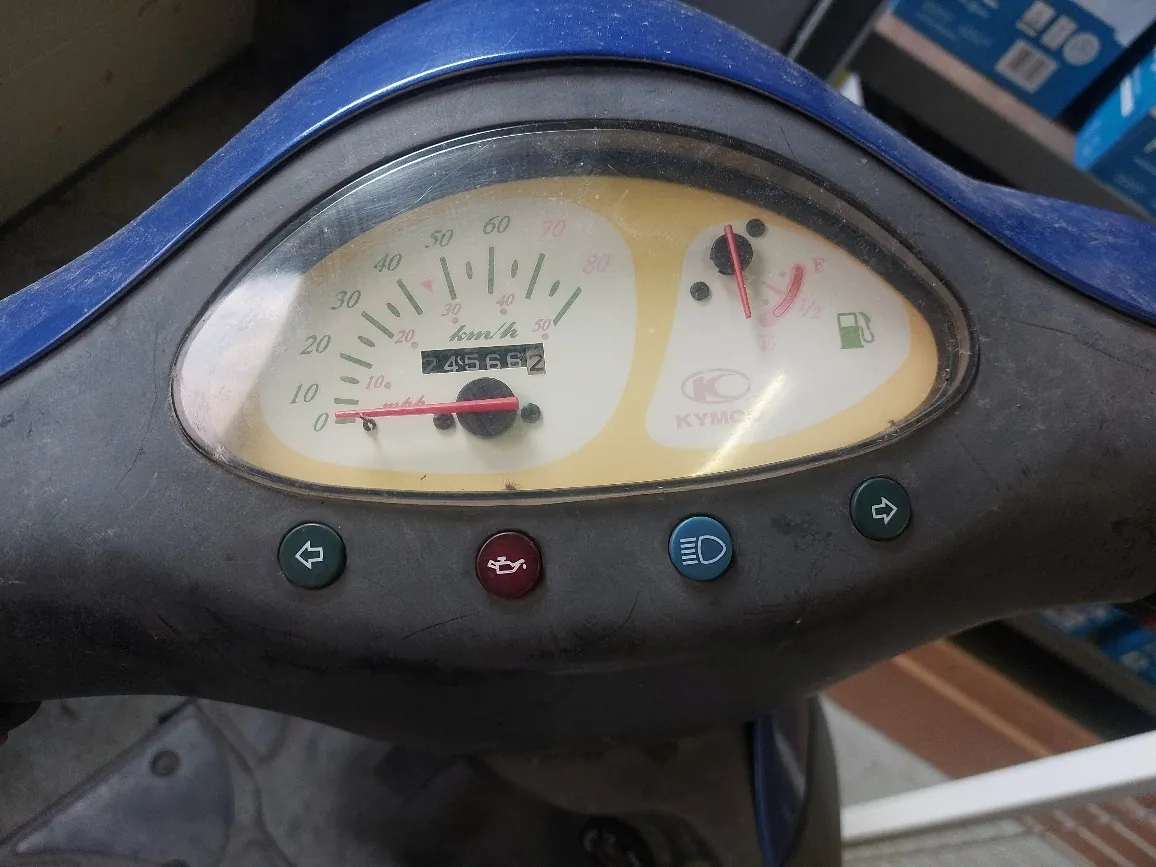 Zbliżenie na deski rozdzielcze skutera marki KYMCO. Widoczny jest prędkościomierz z zakresem do 80 km/h, cyfrowy licznik przebiegu pokazujący 2456,6 km oraz wskaźniki: paliwa, poziomu paliwa (E-F), sygnałów świetlnych i dalekiego światła. Na desce znajdują się cztery przyciski: dwa zielone z ikonami kierunkowskazów, czerwony z ikoną silnika i niebieski z ikoną dalekiego światła. Tło zawiera fragmenty niebieskich pudełek i podłogi.