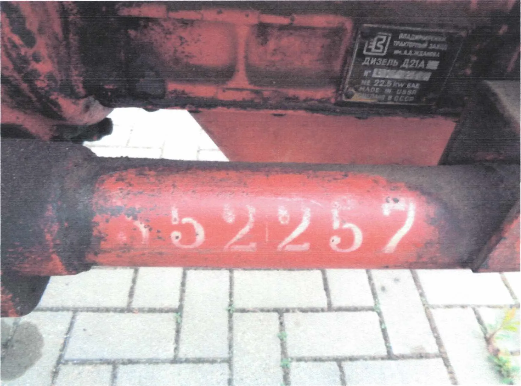 Czerwony metalowy element z białymi cyframi 152257 namalowanymi na powierzchni. W tle metalowa tabliczka z napisem w języku rosyjskim: ВЛАДИМИРСКИЙ ТРАКТОРНЫЙ ЗАВОД им. А.А. ЖДАНОВА ДИЗЕЛЬ Д21А № 152257 22.5 кВт 848 MADE IN USSR СДЕЛАНО В СССР. Podkładka z białych płytek chodnikowych.