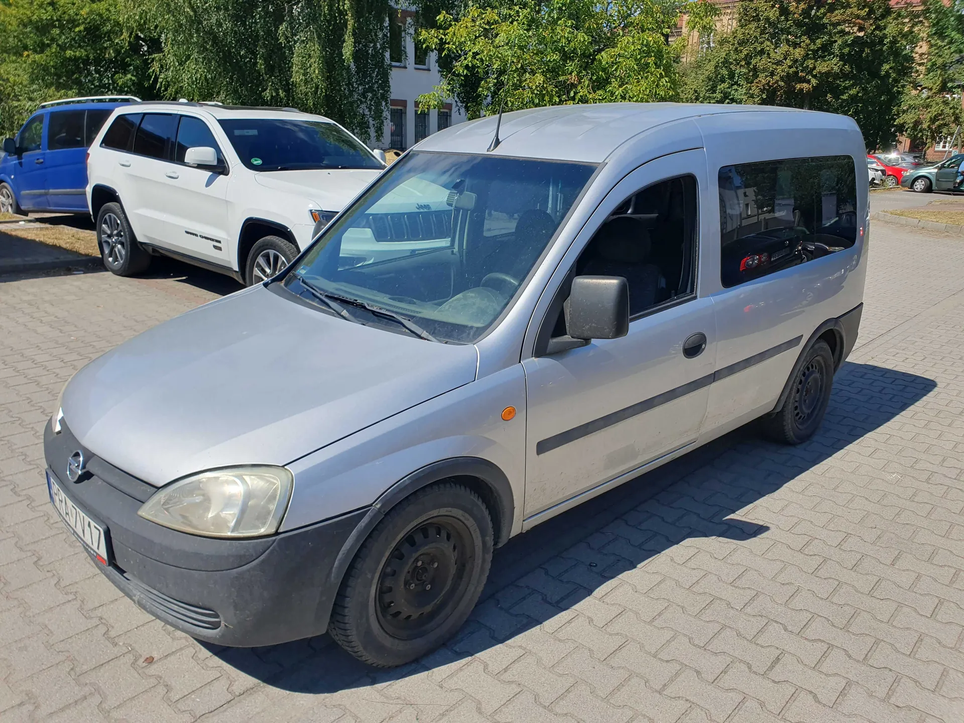Srebrny samochód dostawczy marki Opel Combo zaparkowany na kostce brukowej. W tle widoczny jest biały samochód typu SUV oraz niebieski samochód, a za nimi zielone drzewa i fragment budynku.