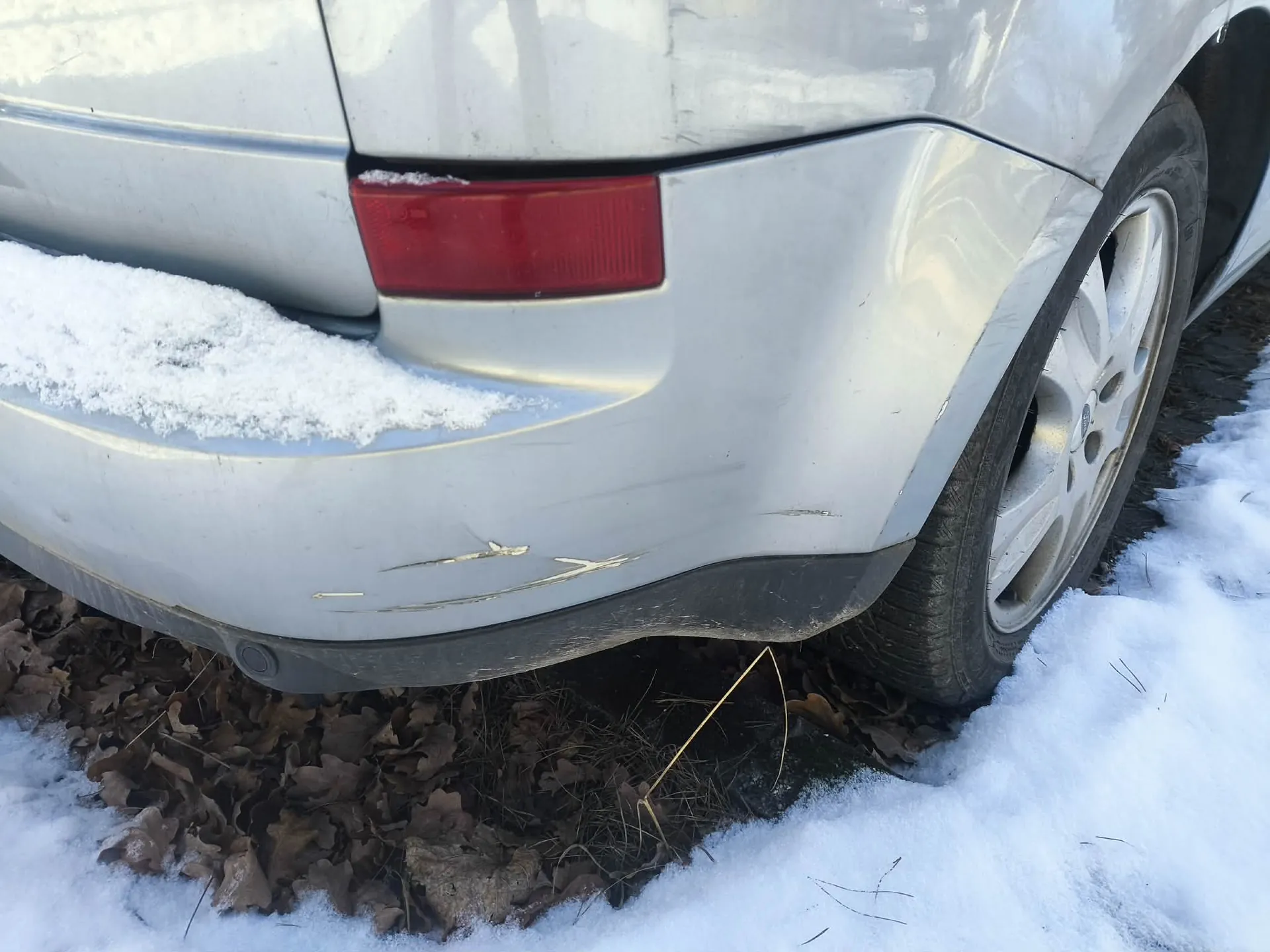 Zbliżenie na tylną część srebrnego samochodu. Widoczny czerwony reflektor, zarysowane zadrapania na zderzaku oraz śnieg na błotniku i ziemi. Pod samochodem suchy liść i trawa wystająca spod śniegu. Opona z aluminiową felgą po prawej stronie.