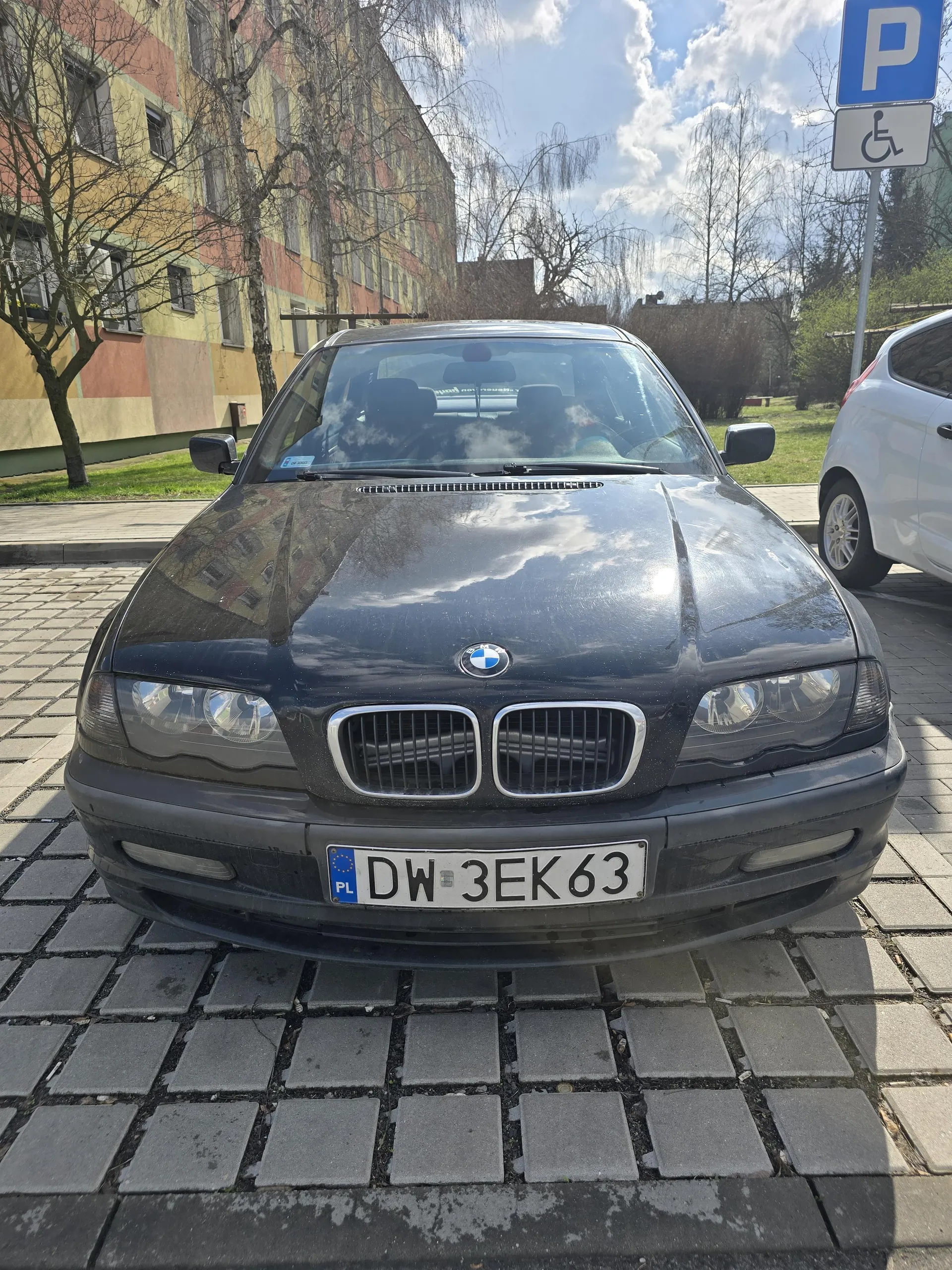 Czarny samochód marki BMW zaparkowany na brukowanej powierzchni. Widoczna tablica rejestracyjna z polskim kodem PL i numerem DW 3EK63. Po prawej stronie biały samochód, a w tle kolorowy blok mieszkalny, drzewa bez liści i niebo z chmurami. Obok samochodu znak parkingowy z literą P oraz symbolem niepełnosprawności.