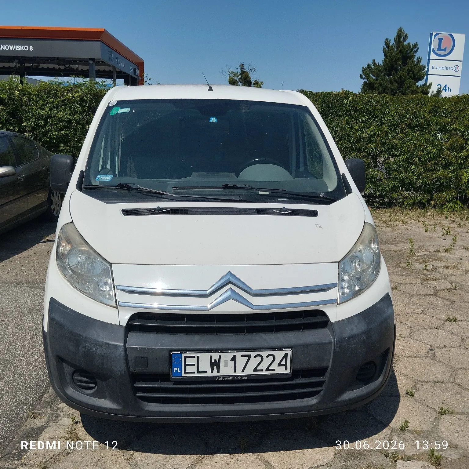 Biały samochód dostawczy marki Citroën zaparkowany na betonowym placu przed zielonym żywopłotem. Pojazd ma polską tablicę rejestracyjną o numerze ELW 17224. W tle widoczny jest fragment budynku z napisem 'NOWISKO 8' oraz niebieska tablica informacyjna z logo i napisem 'E. Lecierc 24h'. Na dole zdjęcia znajduje się znak wodny: 'REDMI NOTE 12' oraz data i godzina '30.06.2026 13:59'.