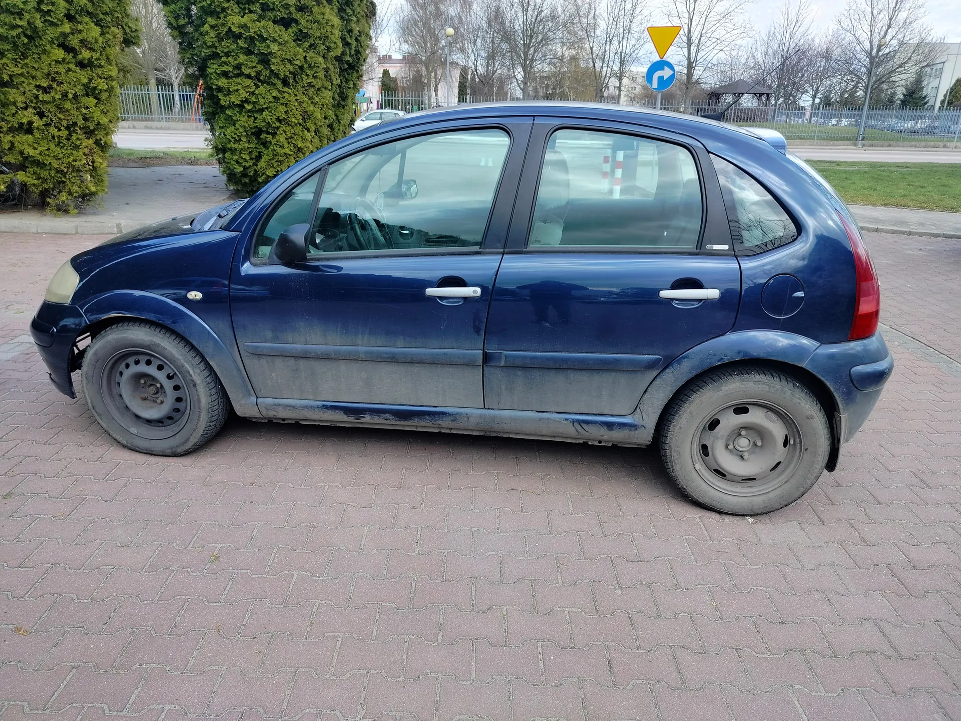 Niebieski samochód osobowy typu hatchback z czterema drzwiami, zaparkowany na brukowanej powierzchni. Pojazd ma zabrudzoną karoserię, szczególnie w dolnej części, oraz felgi stalowe bez osłon. Widoczne uszkodzenie przedniego lewego zderzaka. W tle krzewy, ogrodzenie, drzewa bez liści, drogowskaz z żółtym trójkątem i niebieskim okręgiem z białą strzałką skręcającą w prawo, a także budynki i trawnik.